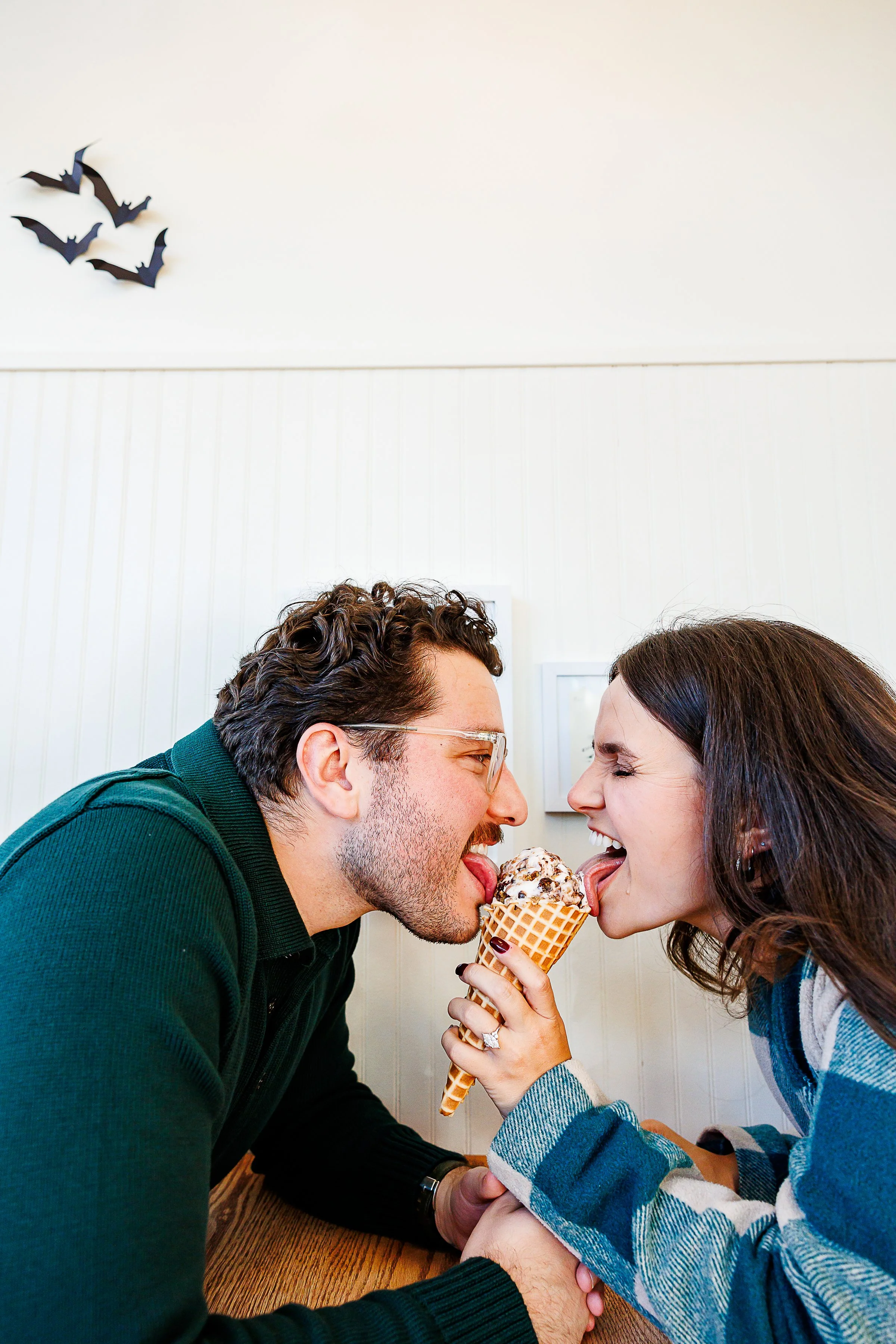 Couple laughing while sharing a bite of an ice cream cone together, her engagement ring visible on her hand holding the cone, playful Halloween bat decor on the wall behind them at Union Market in Richmond, VA.