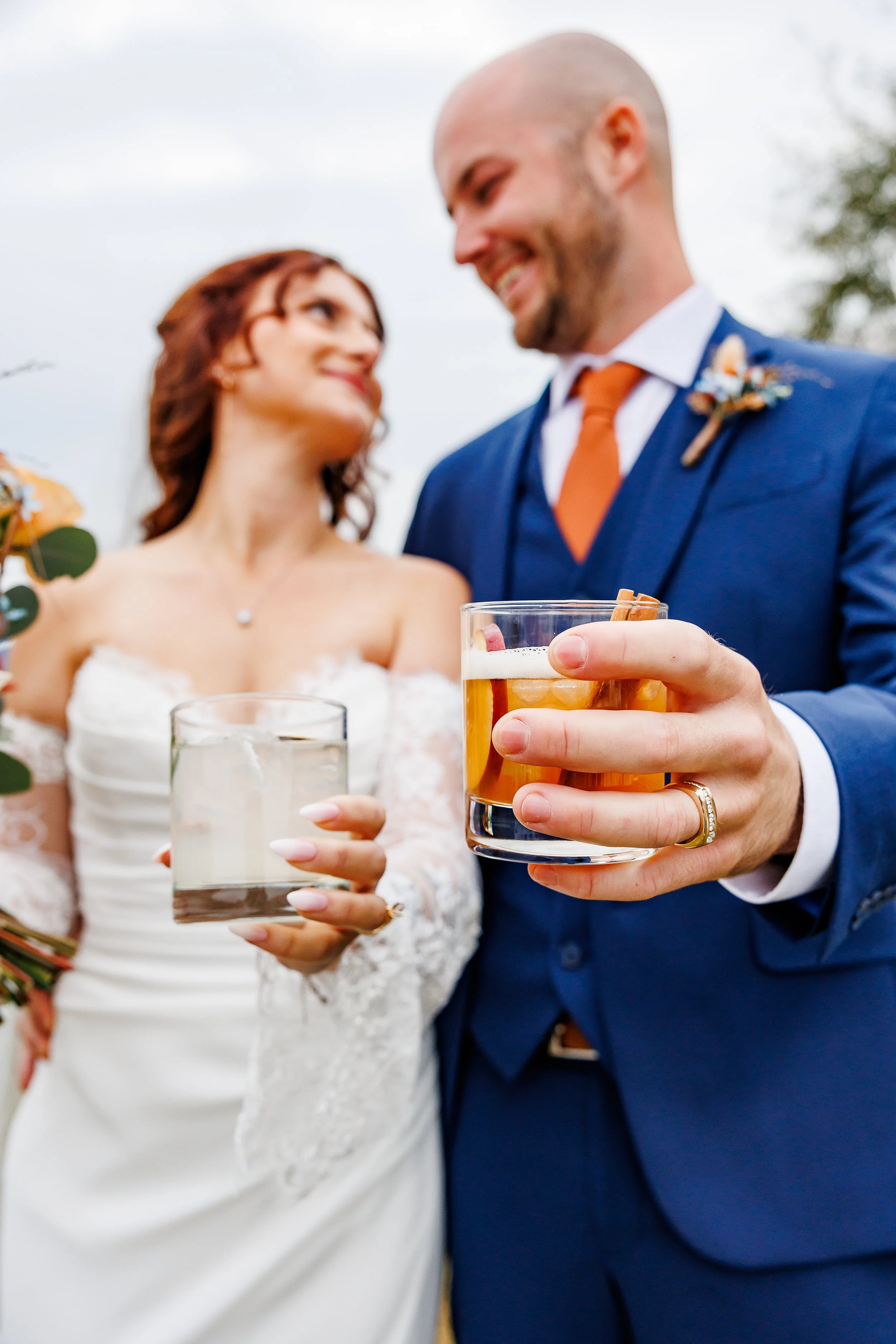 A joyful newlywed couple stands close together, smiling at each other while holding up drinks in a celebratory toast. The groom, in a blue suit with an orange tie, extends an old fashioned toward the camera, while the bride in a lace wedding gown hol