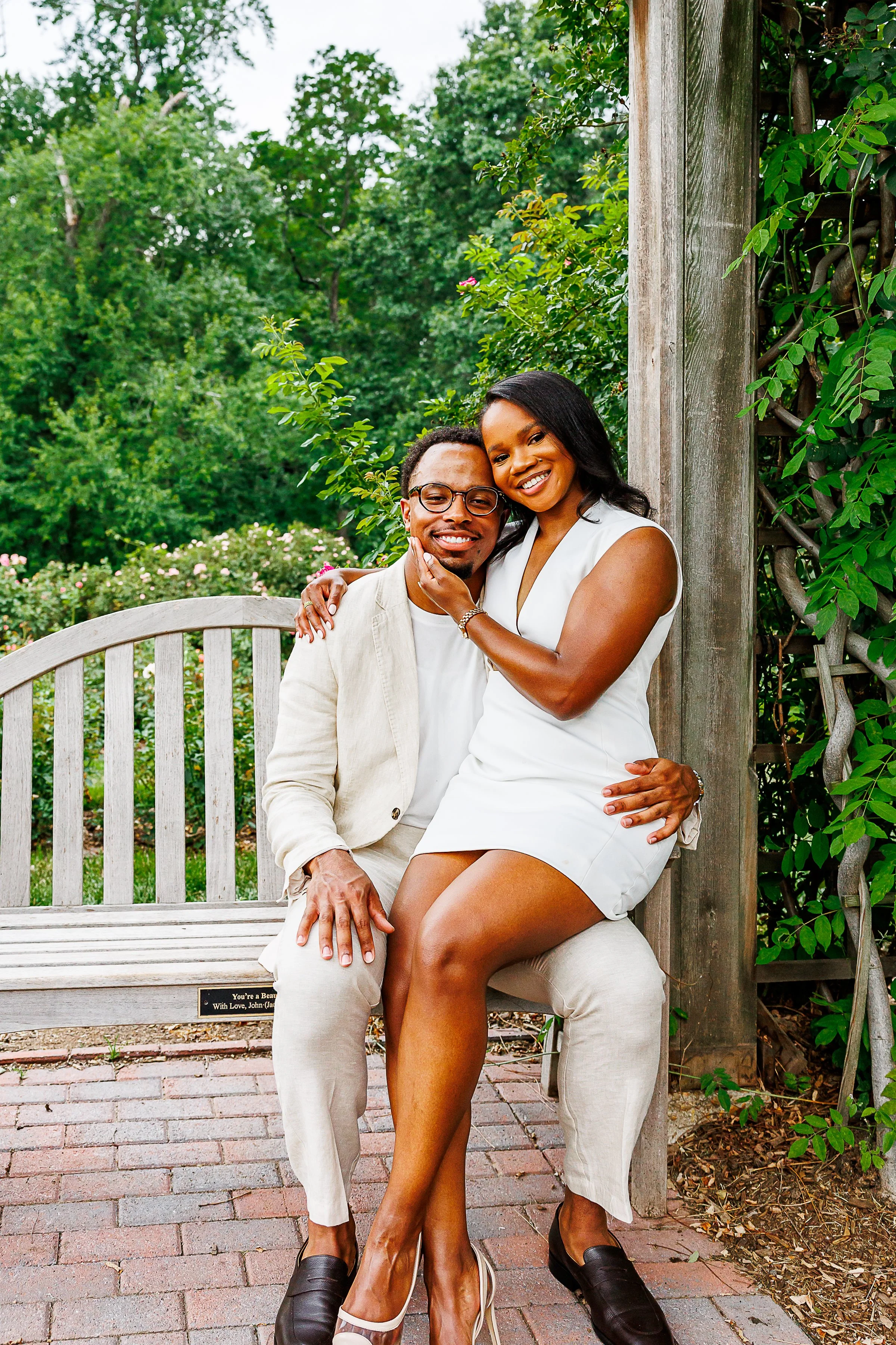 A smiling couple poses together on a wooden garden bench beneath a vine-covered pergola. One partner sits while the other sits sideways on their lap with an arm around their shoulders and a hand gently resting on their cheek. Both wear coordinated li