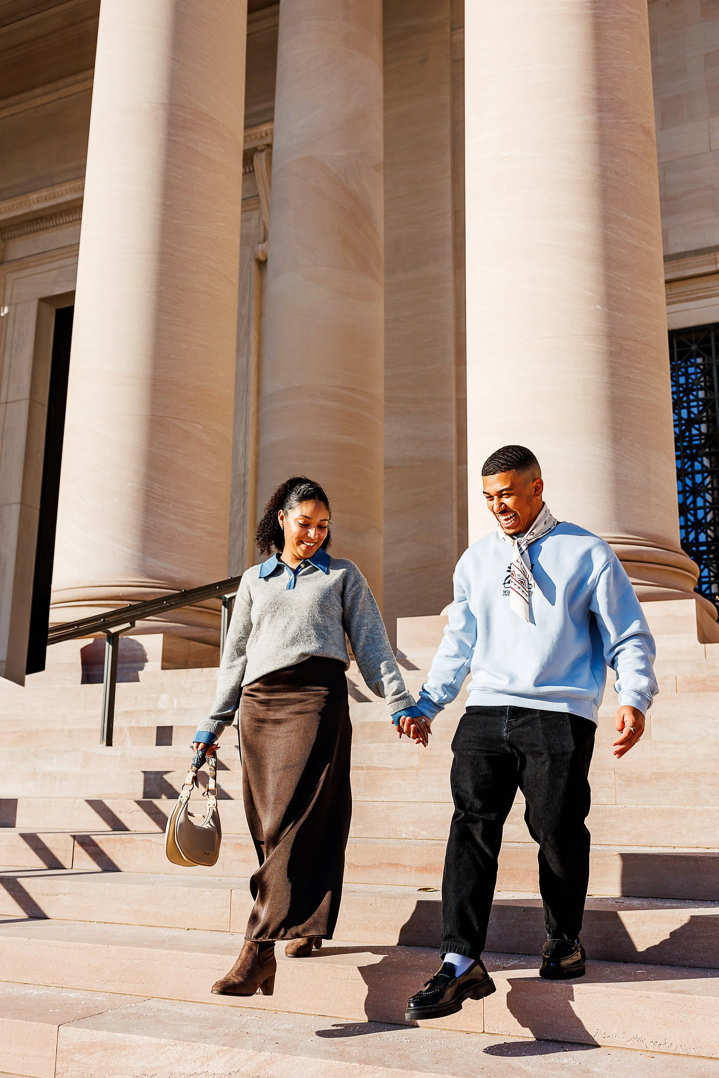Couple holding hands and walking down grand stone steps in front of classical columns in front of the national gallery of art during an outdoor engagement session, smiling in warm natural sunlight.