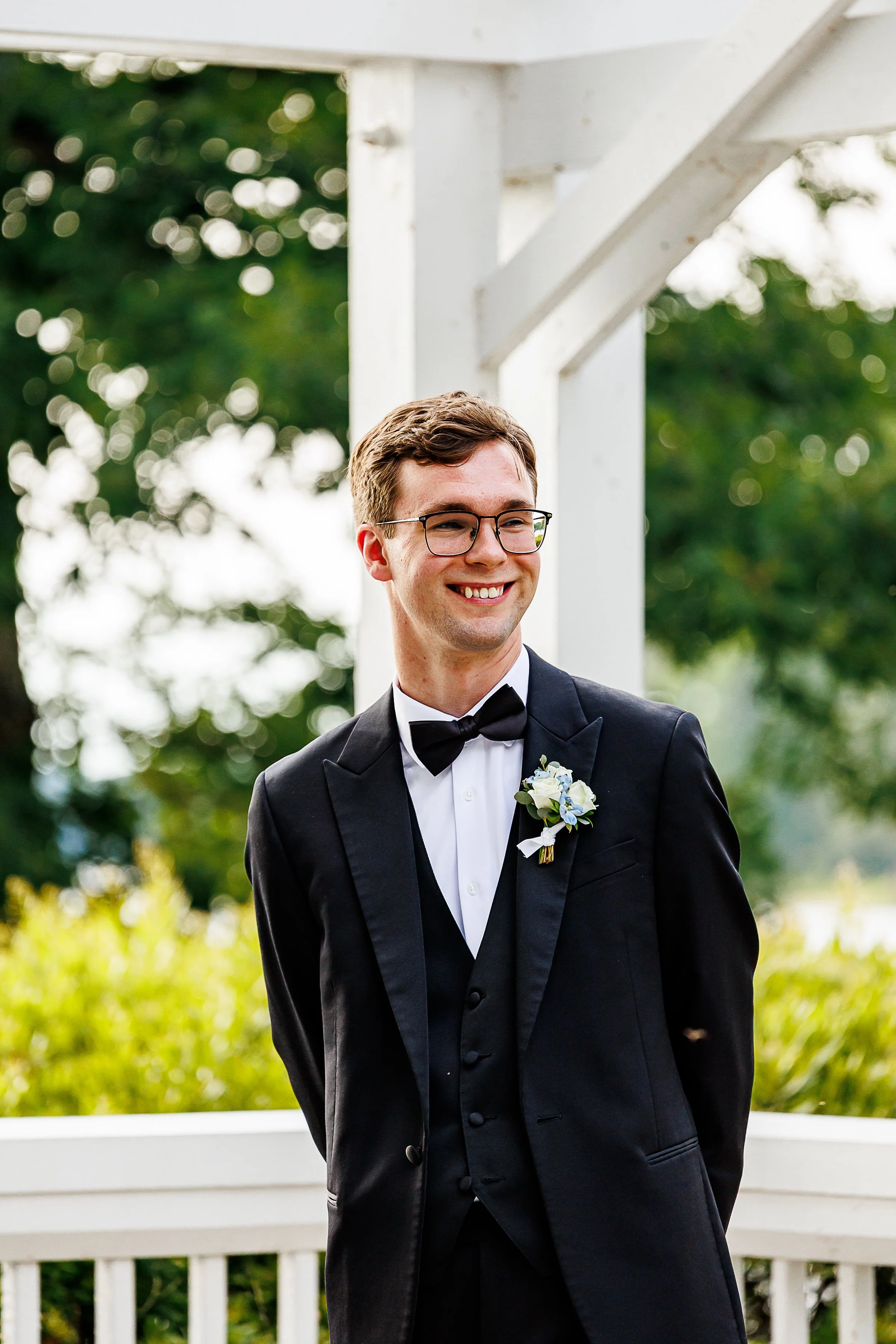 portrait of a smiling groom standing beneath a white waterfront pavilion during an outdoor wedding ceremony. he wears a classic black tuxedo with a bow tie, glasses, and a white floral boutonniere. soft greenery and bright natural light in the backgr