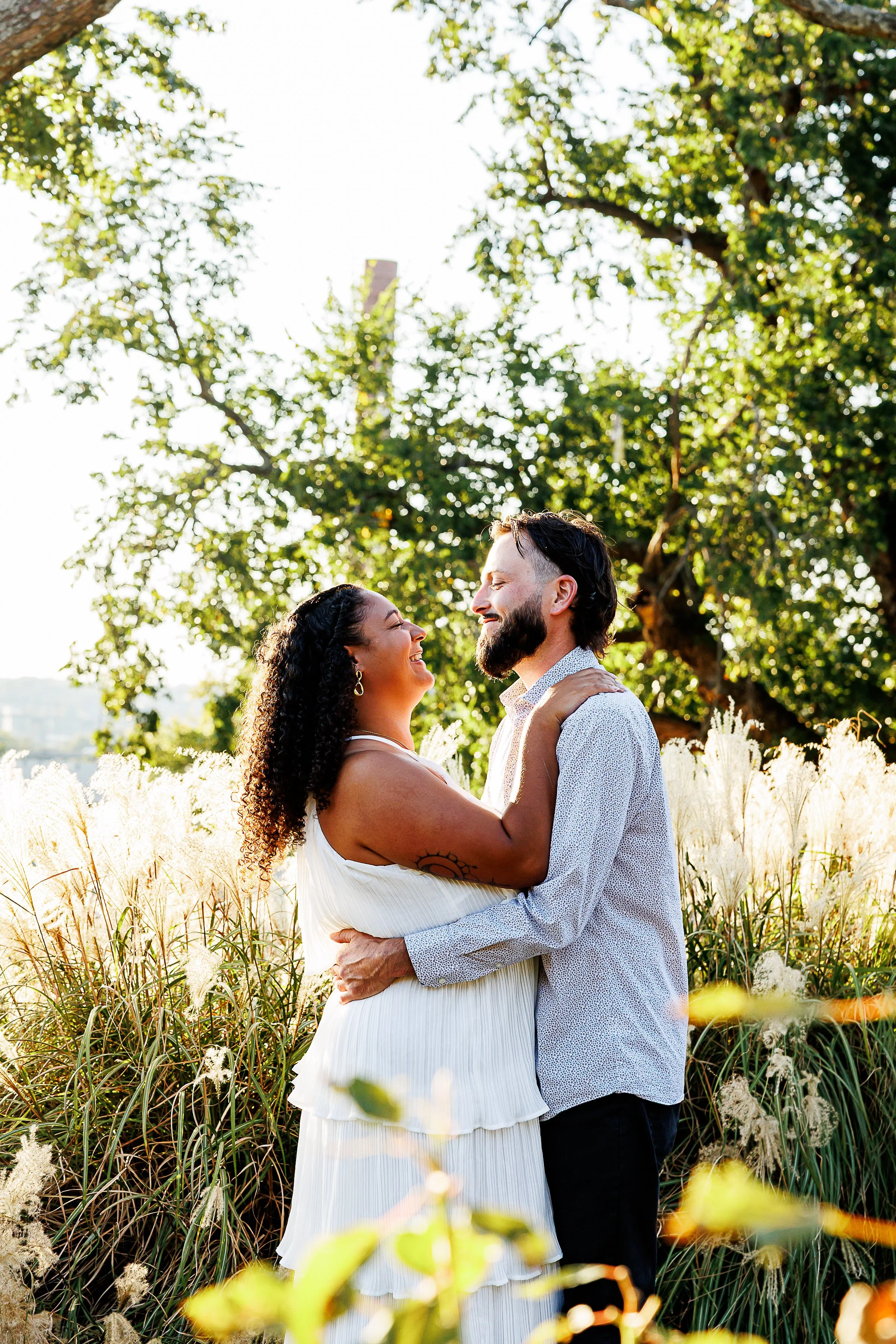 Couple embracing and smiling at each other, her in a white tiered dress with a sun tattoo on her shoulder, surrounded by tall golden grasses at sunset at Libby Hill Park in Richmond, VA.