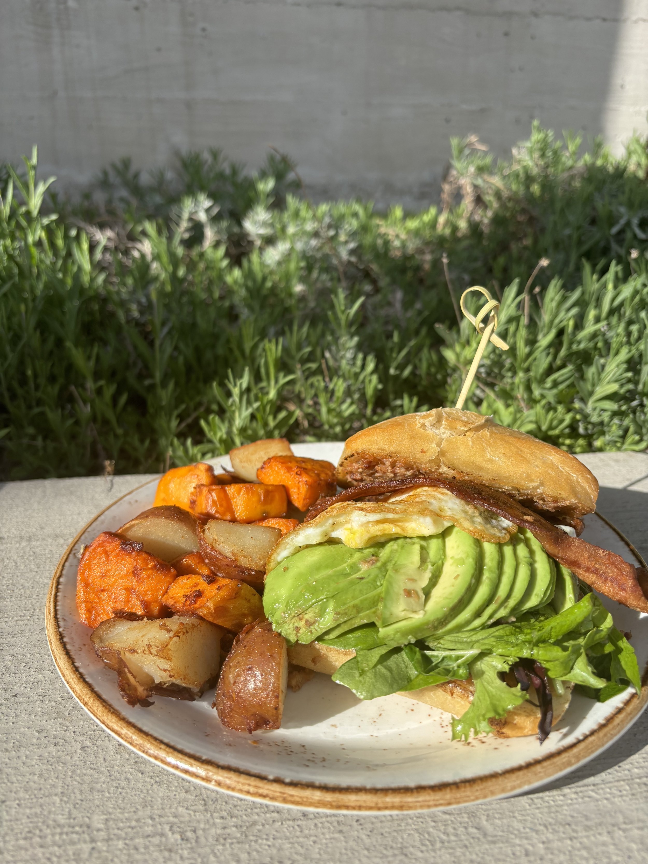Bacon on Bacon Breakfast Sandwich:
A soft ciabatta roll layered with house made bacon onion jam, slices of thick cut bacon, a fried egg, avocado, and citrus dressed greens, all served with a side of our potato medley. A bacon lovers dream!