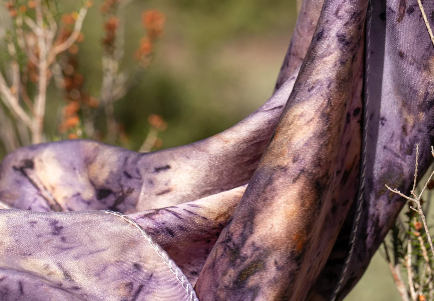 A close-up of a silk scarf, naturally dyed with logwood bark, hibiscus petals and marigold flowers, sitting among dry plants and twigs.