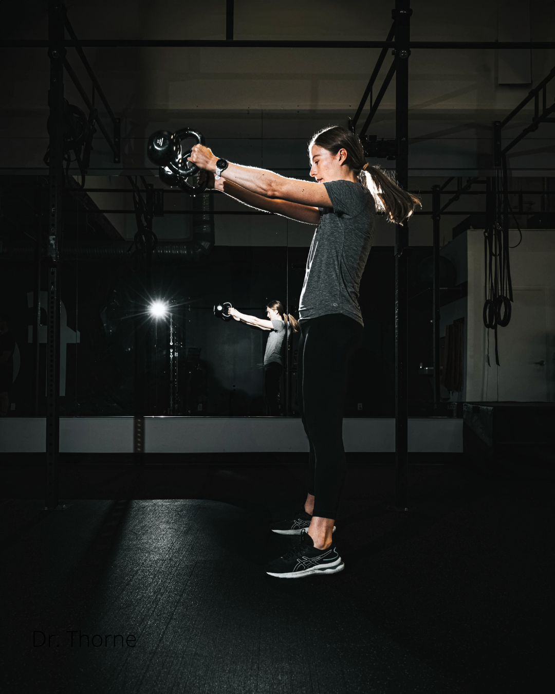 Woman in a gym standing on black rubber flooring, performing a kettlebell exercise with her arms extended forward, with a mirror reflecting her image and gym equipment in the background.