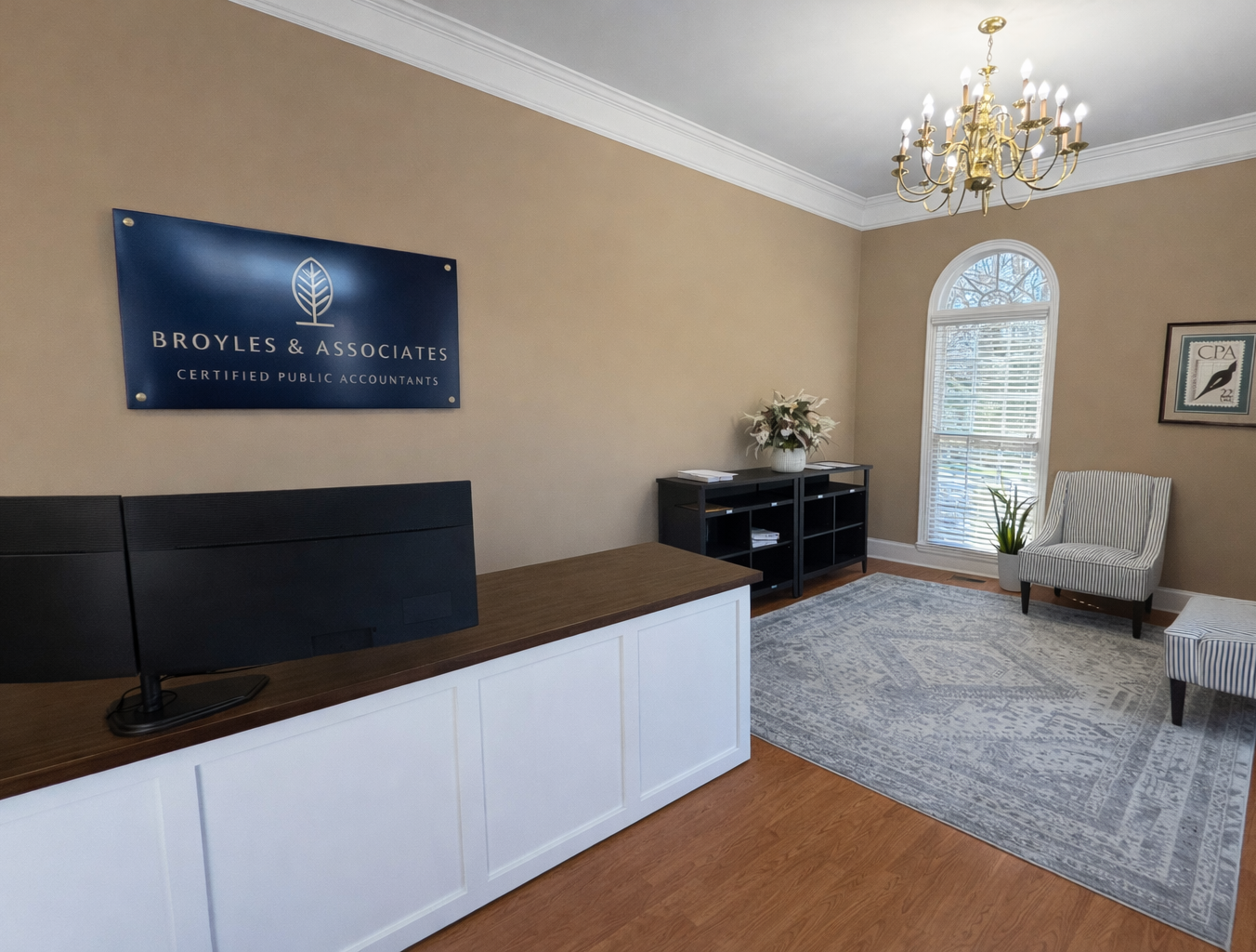 Office reception area with a sign for Broyles & Associates, certified public accountants, a reception desk with dual monitors, a black bookshelf with files, and seating with a potted plant near a window.