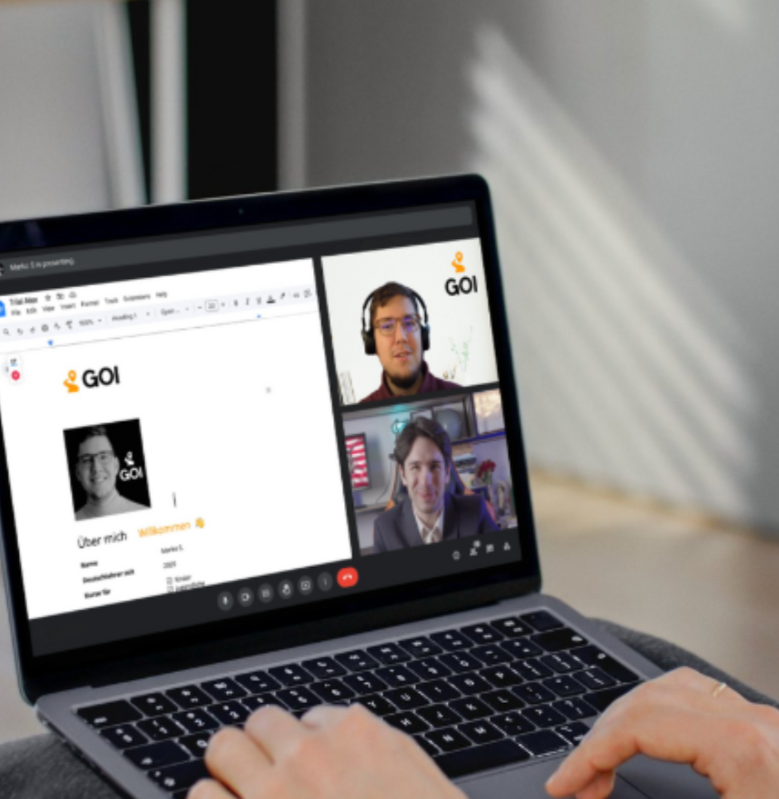 A person using a laptop during a virtual meeting with two other participants, showing a presentation slide with the GOI logo and a black-and-white profile photo.