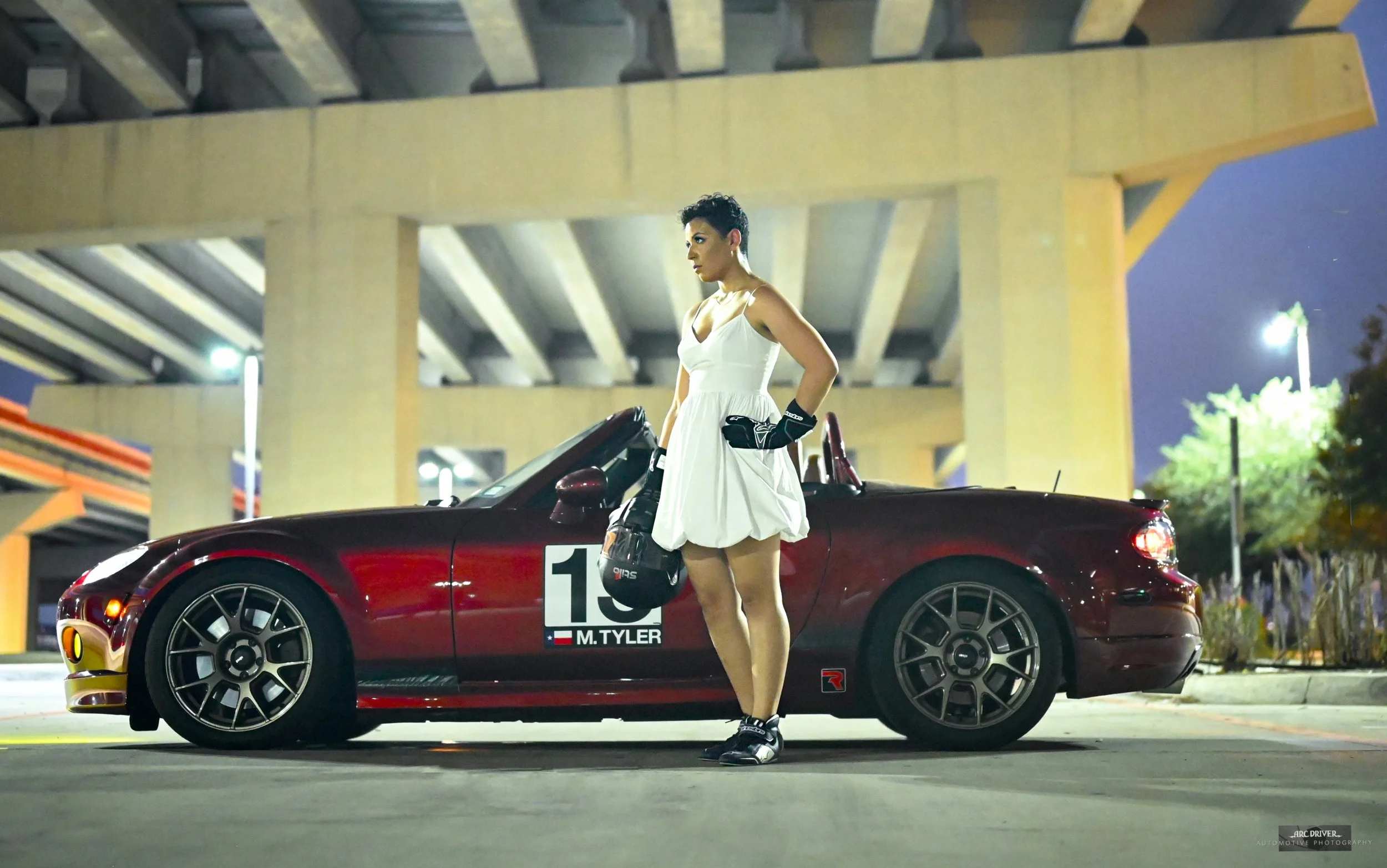 A woman in a white dress and black racing gloves standing next to a red convertible sports car with racing decals, including the name M. Tyler, parked under a concrete highway overpass at night.