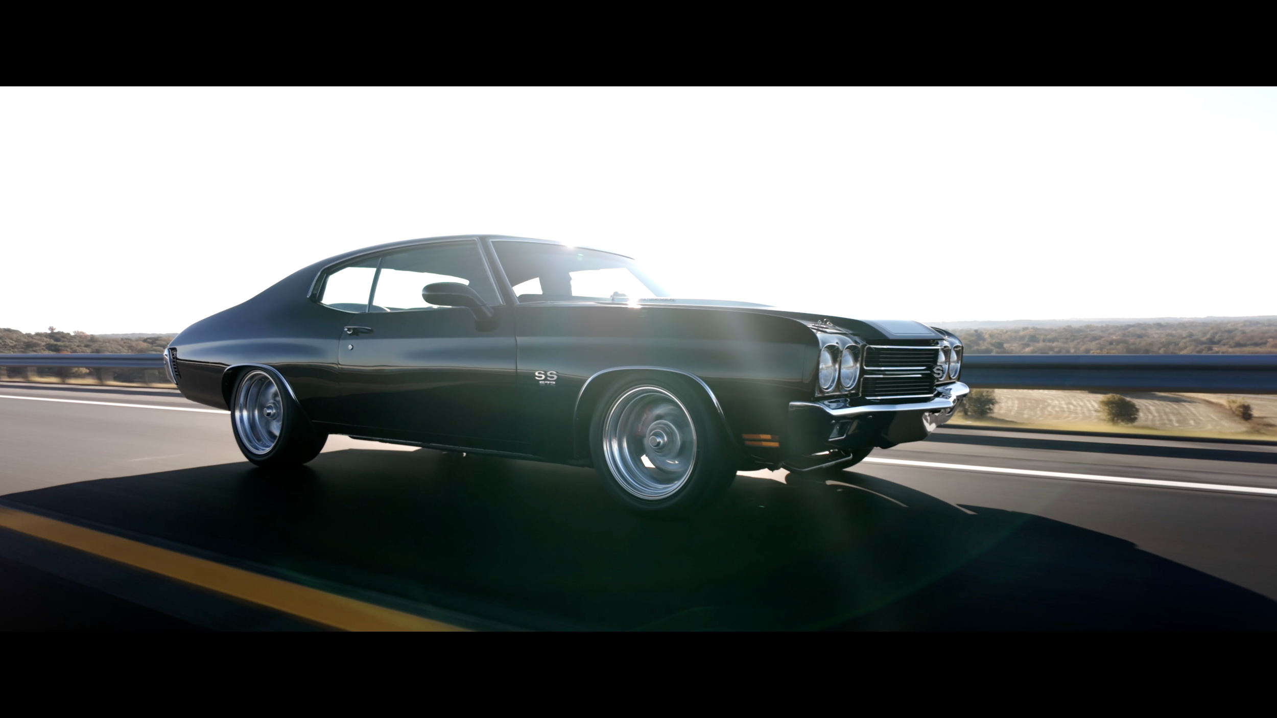 A black classic Chevrolet Chevelle SS muscle car driving on a highway with a scenic landscape in the background during daytime