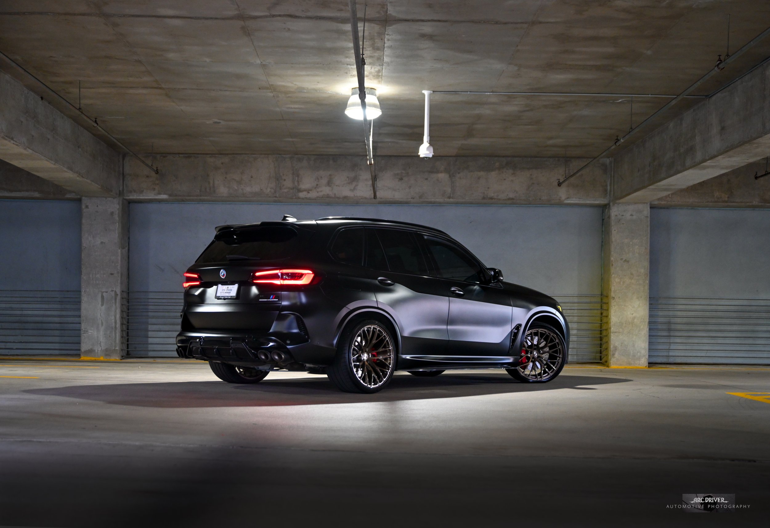 A black high-performance car parked in an empty underground parking garage with concrete walls and ceiling, illuminated by overhead lights.