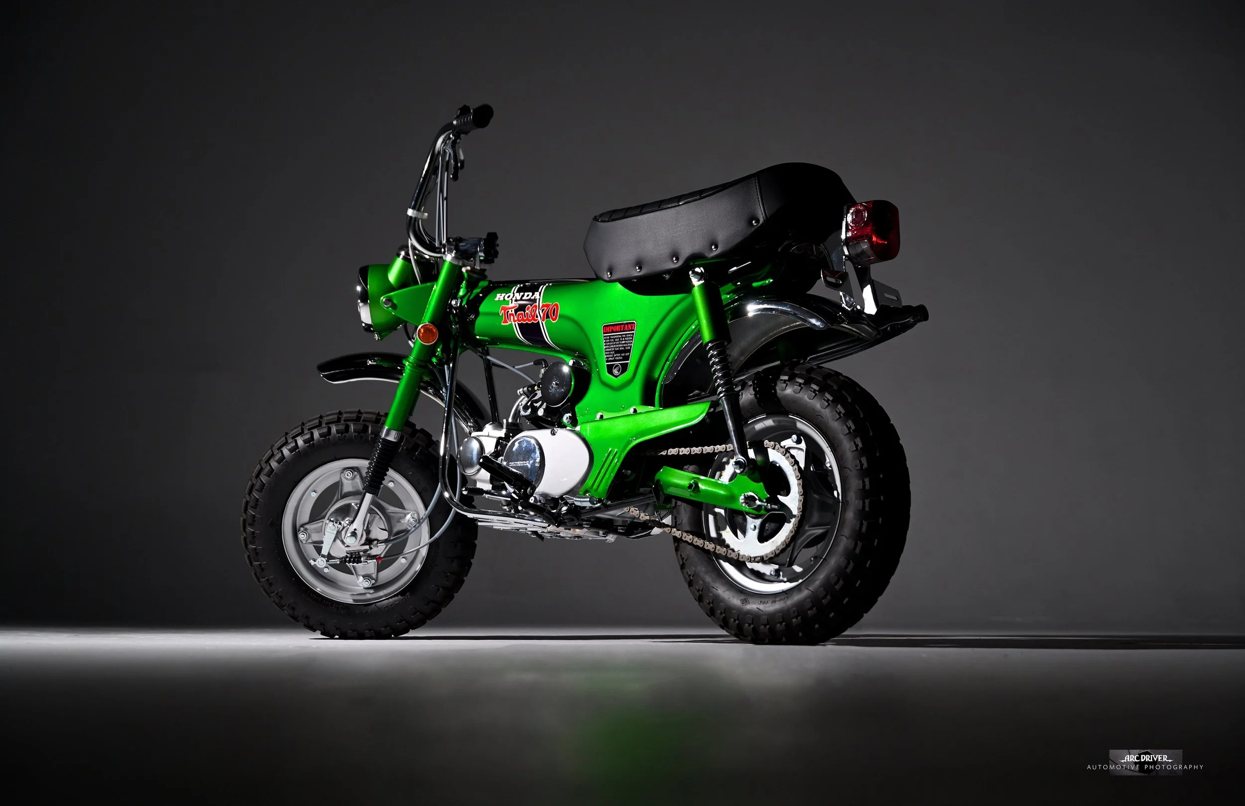 A small green Honda Trail 70 dirt bike with a black seat, positioned against a dark background, illuminated by studio lighting.