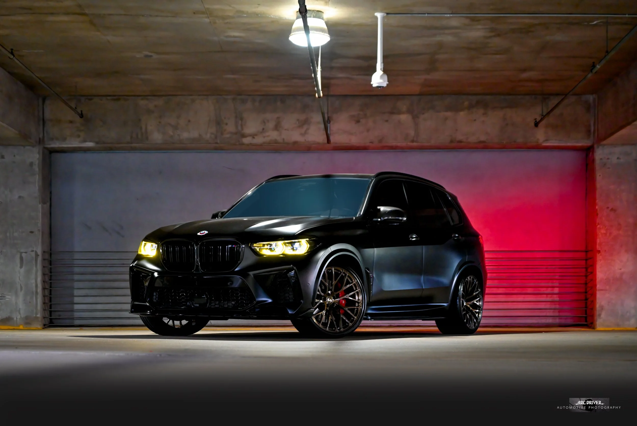 A sleek black luxury SUV with tinted windows and modern headlights, parked in an indoor garage with a concrete and wooden ceiling, illuminated by a ceiling light and red accent lighting.