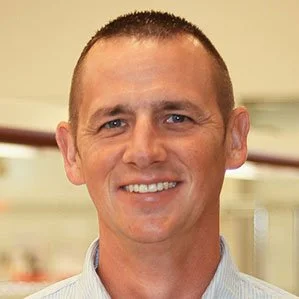 Smiling man with short hair, wearing a striped shirt, in an indoor setting.