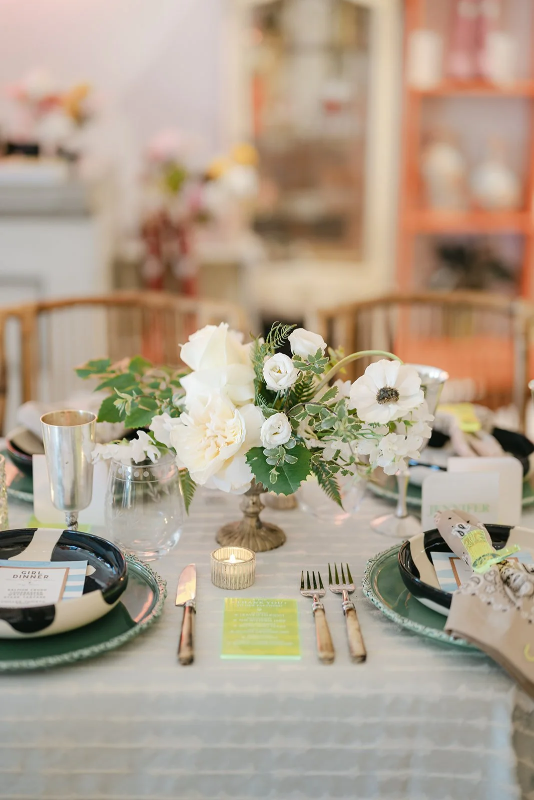 Table decorated with a floral centerpiece of white flowers and green leaves, set with black and white dishes, gold utensils, a candle, and place cards, in a well-lit room.