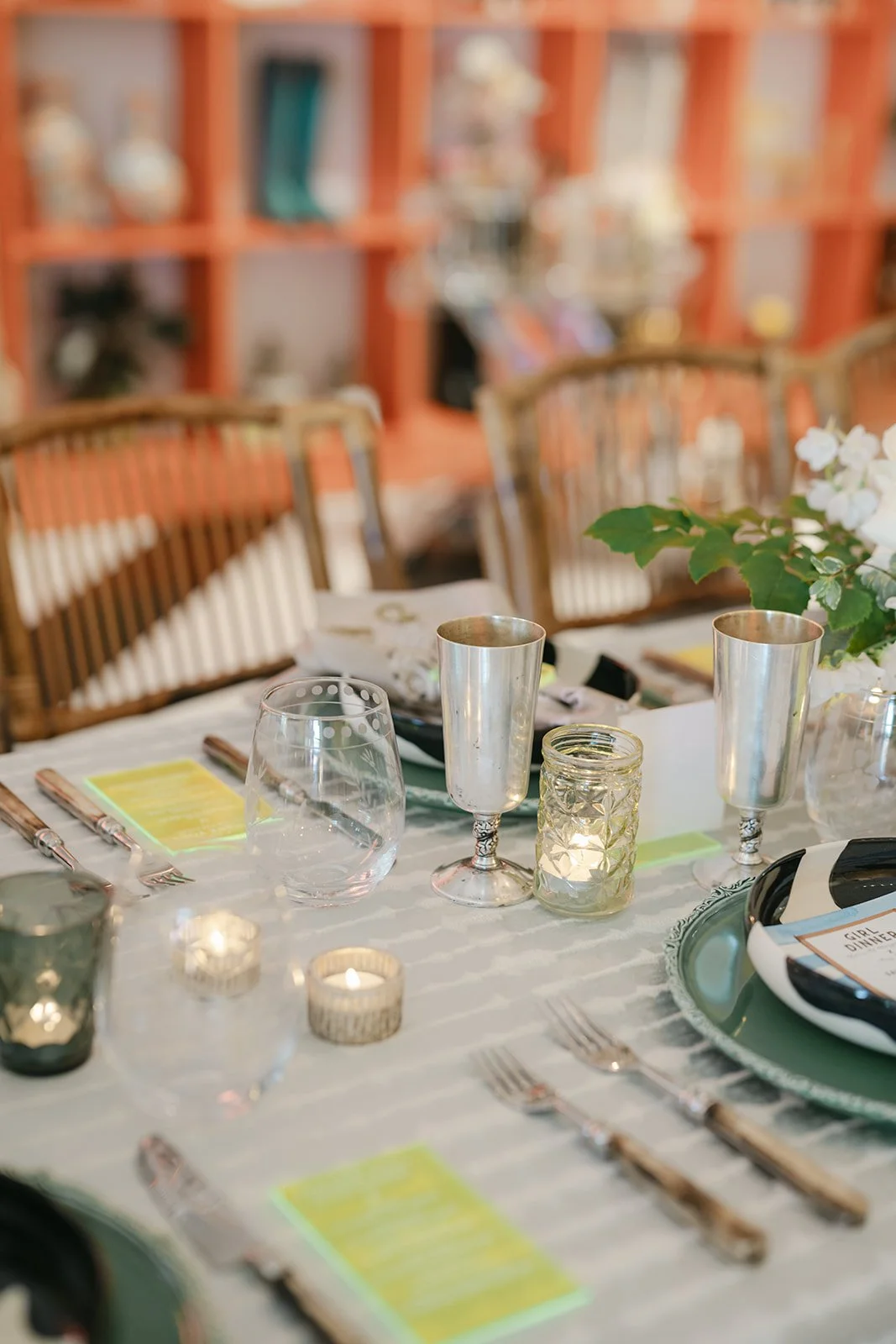 A decorated dining table set with wine glasses, candles, plates with name tags, silverware, and a floral centerpiece, with a blurry background of chairs and a bookshelf.