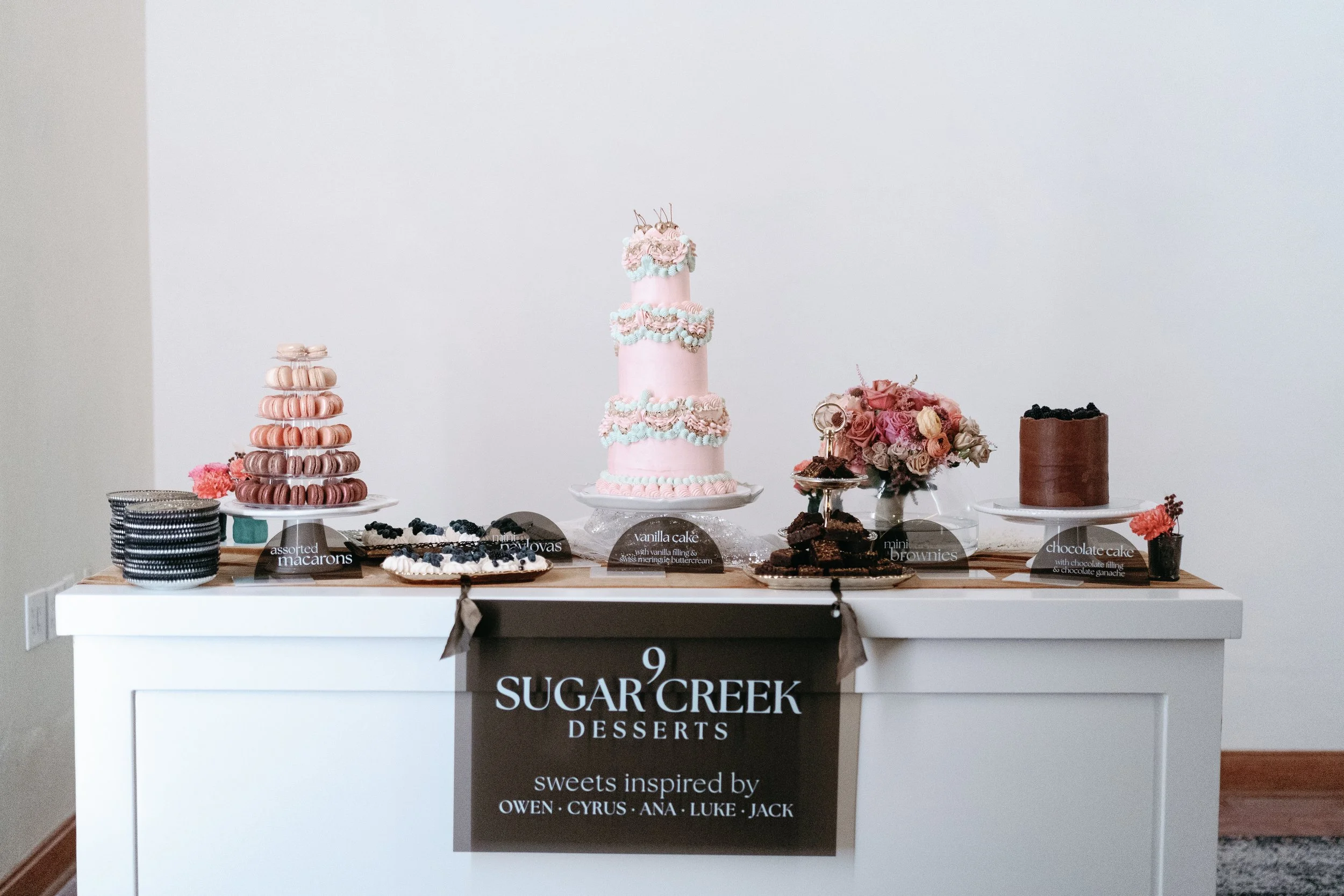 Display table of desserts including a tall pink cake with pastel-colored icing decorations, a stacked macaron tower in shades of pink, beige, and brown, a chocolate cake with berries, and assorted smaller desserts with labels, behind is a plain white