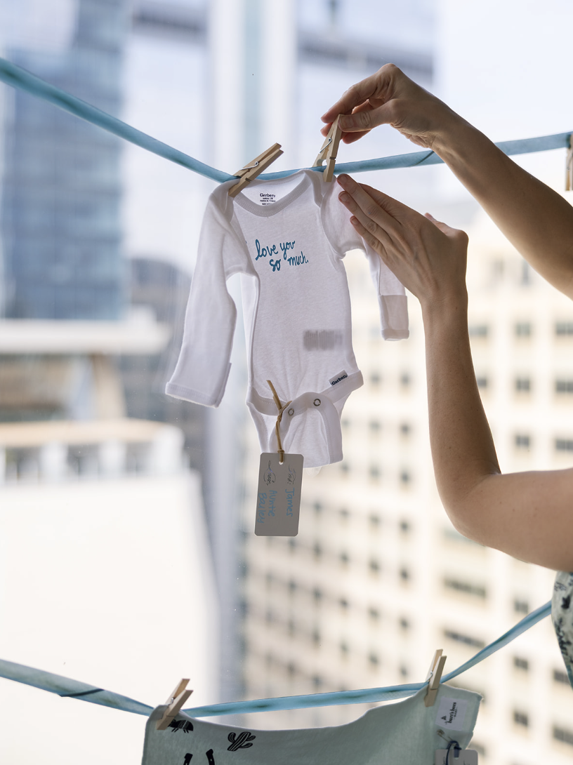Small white baby onesie with blue embroidery hanging on a clothesline, with a background of city buildings visible through a window.