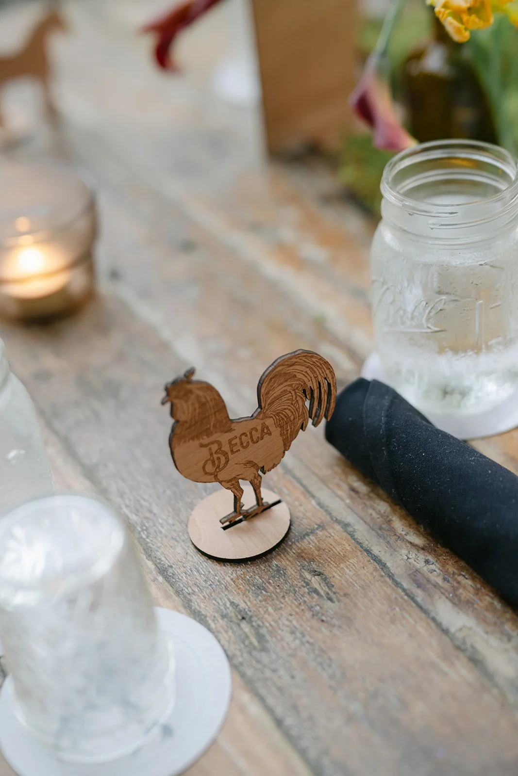 Wooden rooster figurine with the word "BECCA" etched on it, on a rustic wooden table next to a mason jar filled with water.