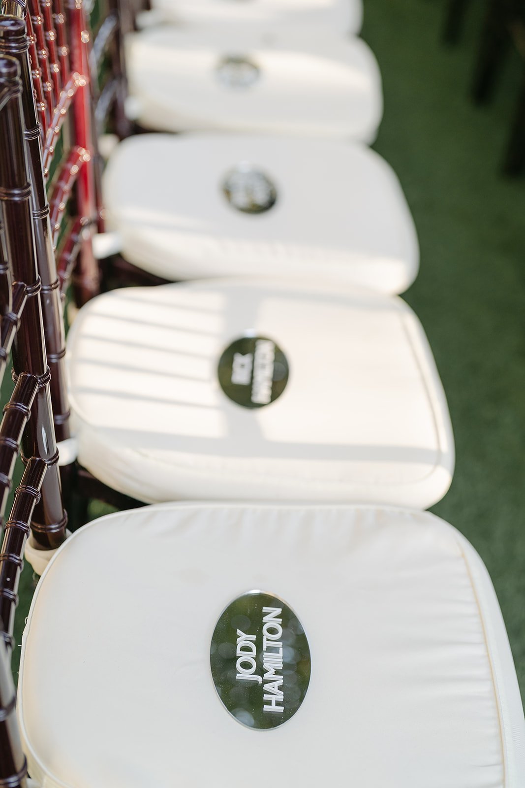 A row of white chairs with Jody Hamilton stickers on the backrests, aligned on a green grassy area, with shadows from nearby structure falling across the chairs.