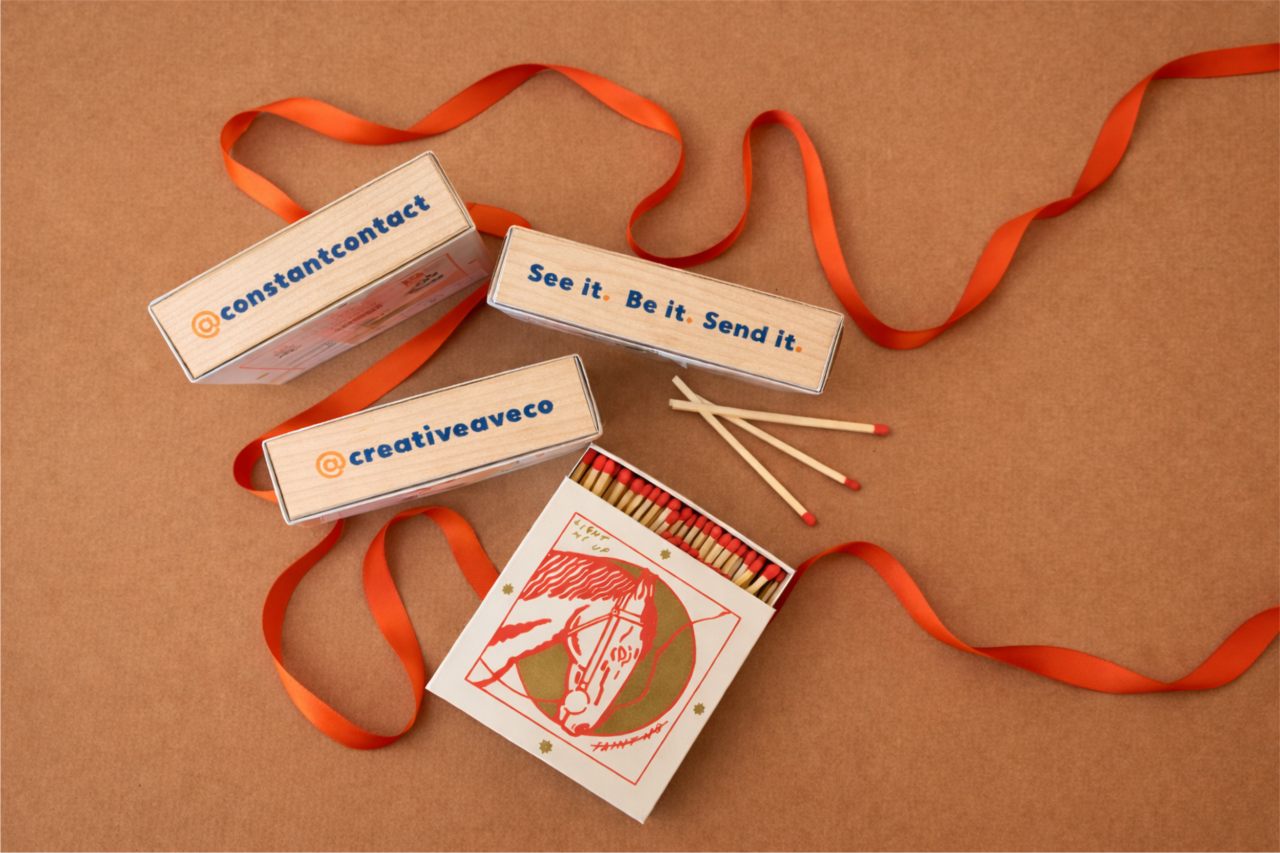 Multiple matchstick boxes with social media handles and slogans, along with some matchsticks, a red ribbon, and a card with an illustration of a person, all arranged on a brown background.