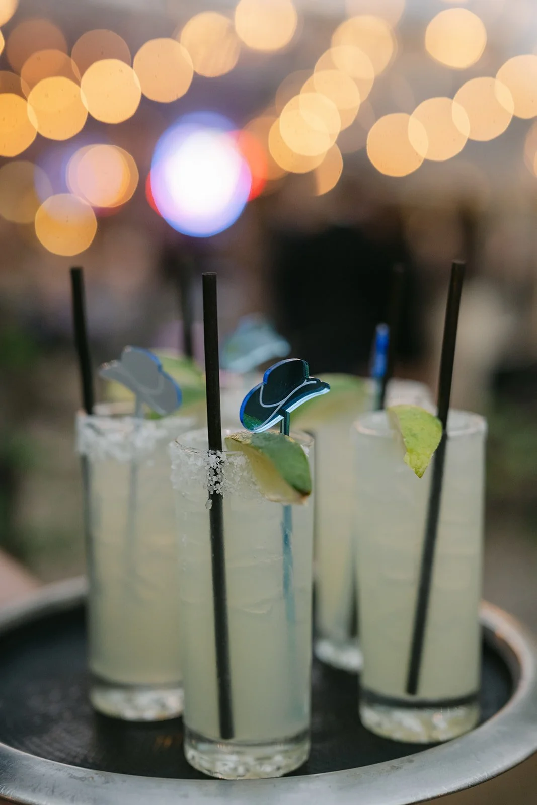 Four glasses of margaritas garnished with lime wedges and small cowboy hat stirrers, with a salt rim on one glass, set against a blurry background with yellow bokeh lights.