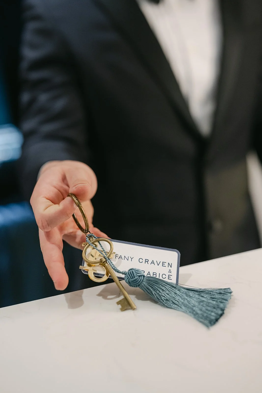 Person in black blazer holding a keychain with a tassel and a card that reads 'Tiffany Craven & Grace'.
