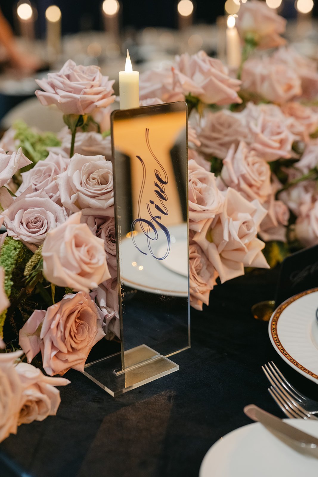 A floral centerpiece of pink roses on a black tablecloth at a formal event with a table number sign and candle.
