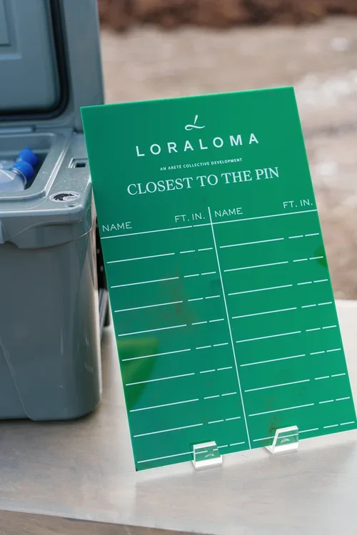 Loraloma sign titled 'Closest to the Pin' with spaces to write names and distances in feet, placed on a table near a cooler in an outdoor setting.