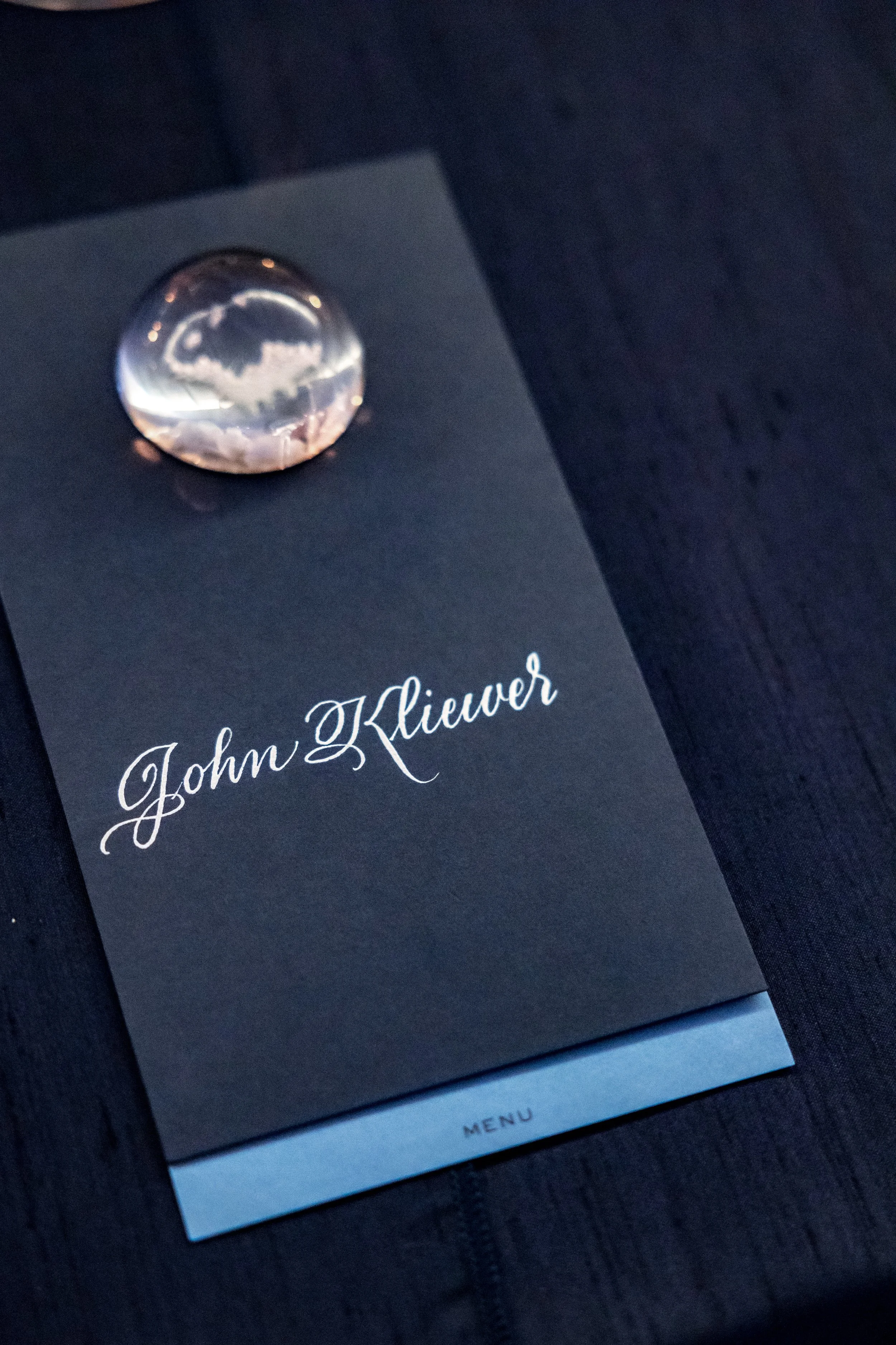 Close-up of a menu titled 'John Kliever' with a decorative glass ball on top, reflecting the surrounding environment, placed on a dark wooden surface.
