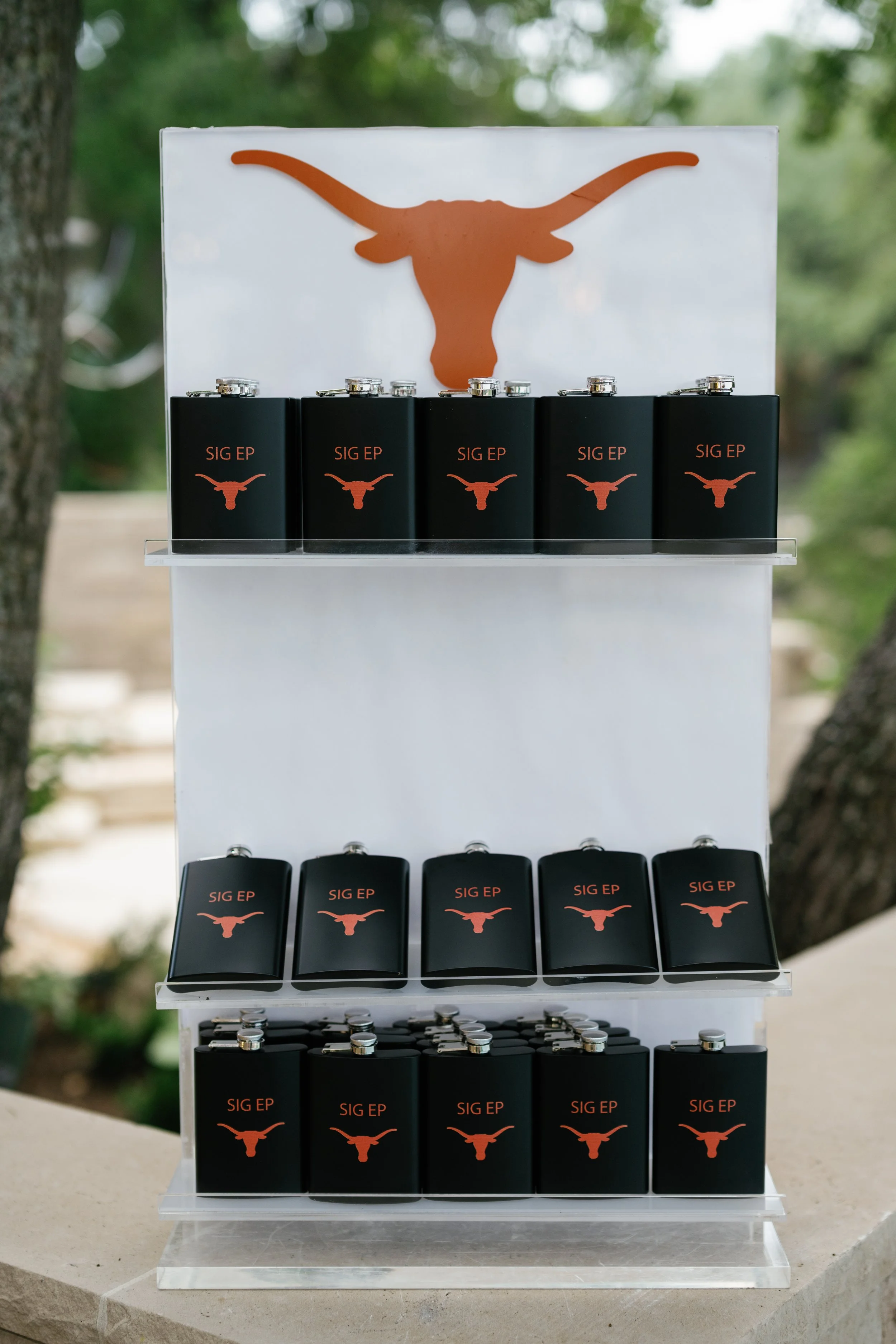 Display of black flasks and Lip balm tubes with a longhorn logo and text 'SIG EP' on a three-tiered white stand outdoors.