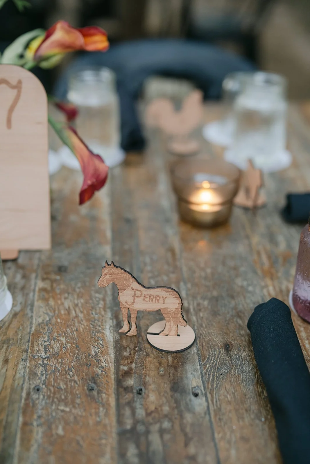 Wooden table with a small wooden horse figurine engraved with the name 'PERRY' in the foreground. In the background, there are candles, jars, and a black object, with a blurred floral arrangement on the left.