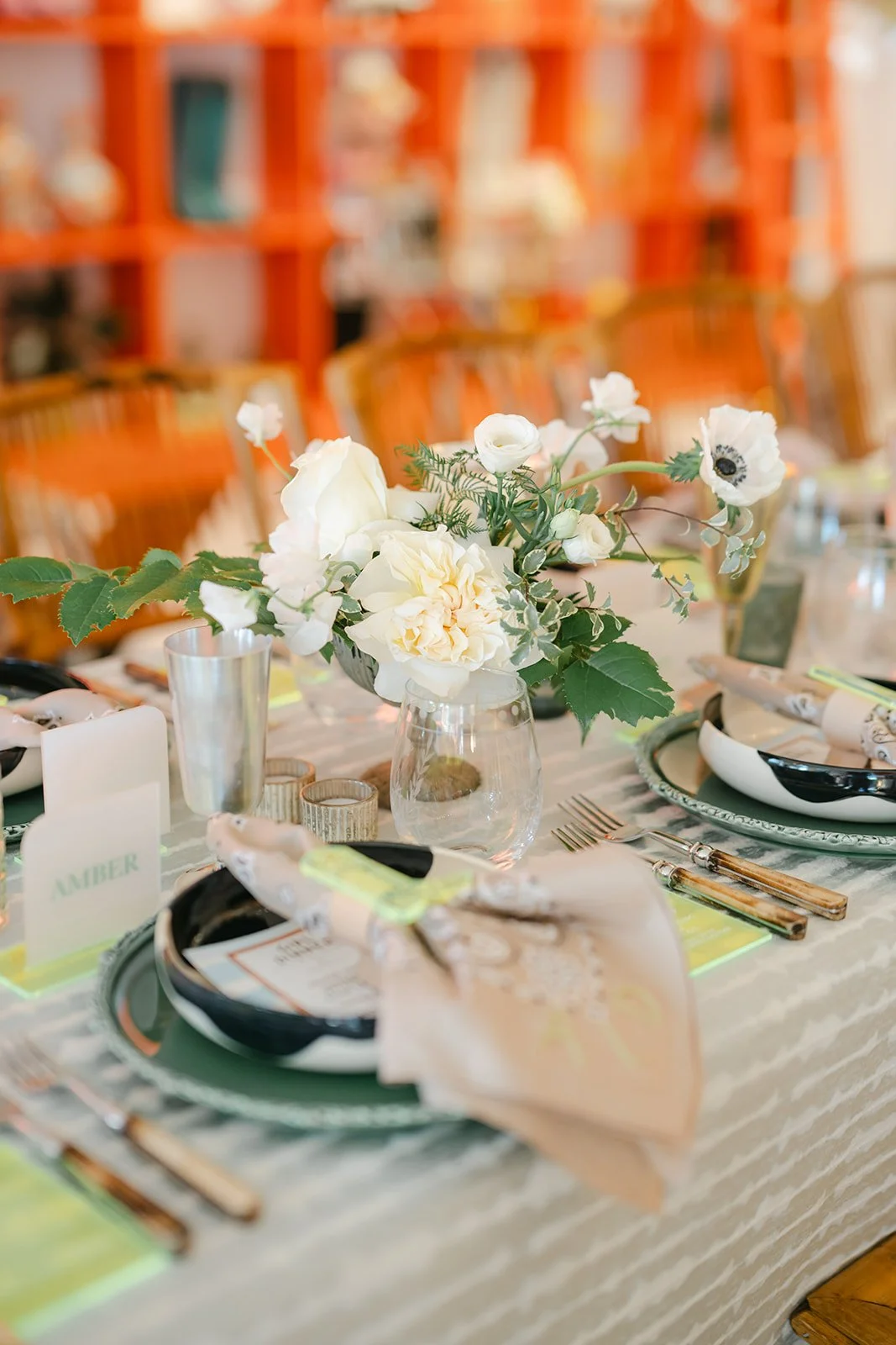 A decorated table with a floral centerpiece, tableware, and name cards for a formal event, in a room with orange shelving in the background.