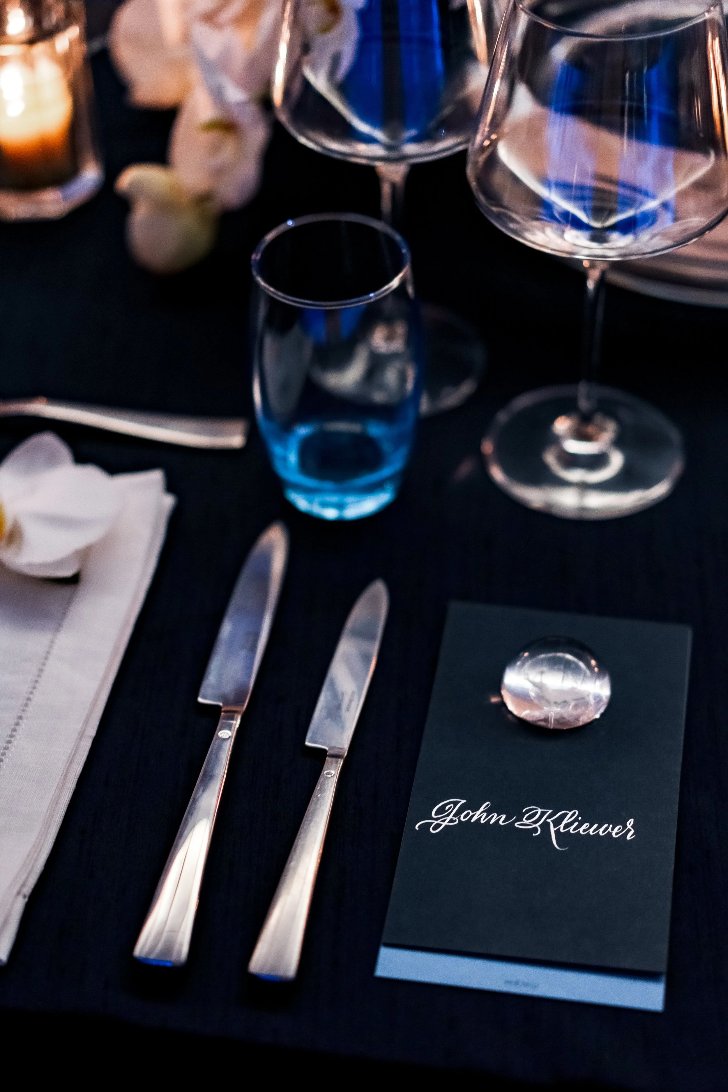 A table setting with a black tablecloth includes a white cloth napkin, a small spoon, a butter knife, a dinner knife, and a dinner fork arranged from left to right. There are two wine glasses and a small water glass, along with a lit candle, flowers,