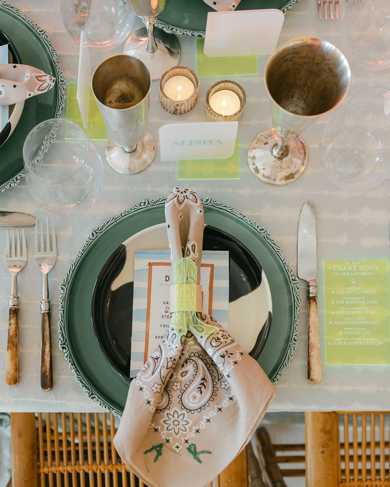 A formal dinner table setting with a green and black patterned charger plate, a menu, white napkin with a paisley pattern, and silverware. There are candles and empty wine glasses on the table. A name card with "ALISHA" is visible.