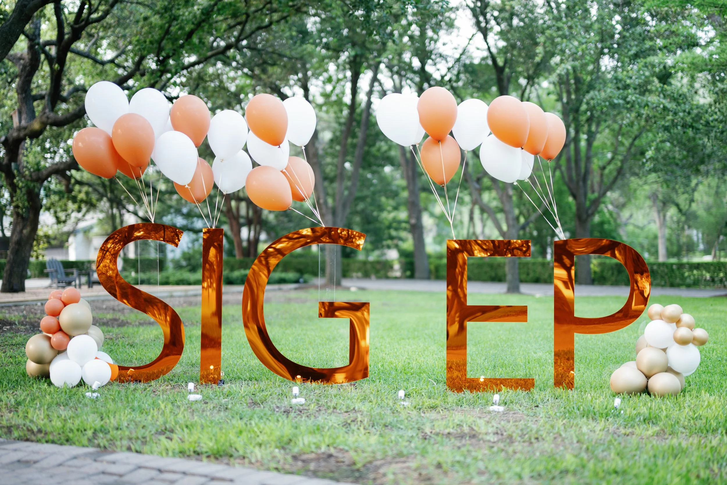 Outdoor celebration setup with orange and white balloons attached to the top of large, metallic copper-colored G and E letters on a grassy area surrounded by trees.