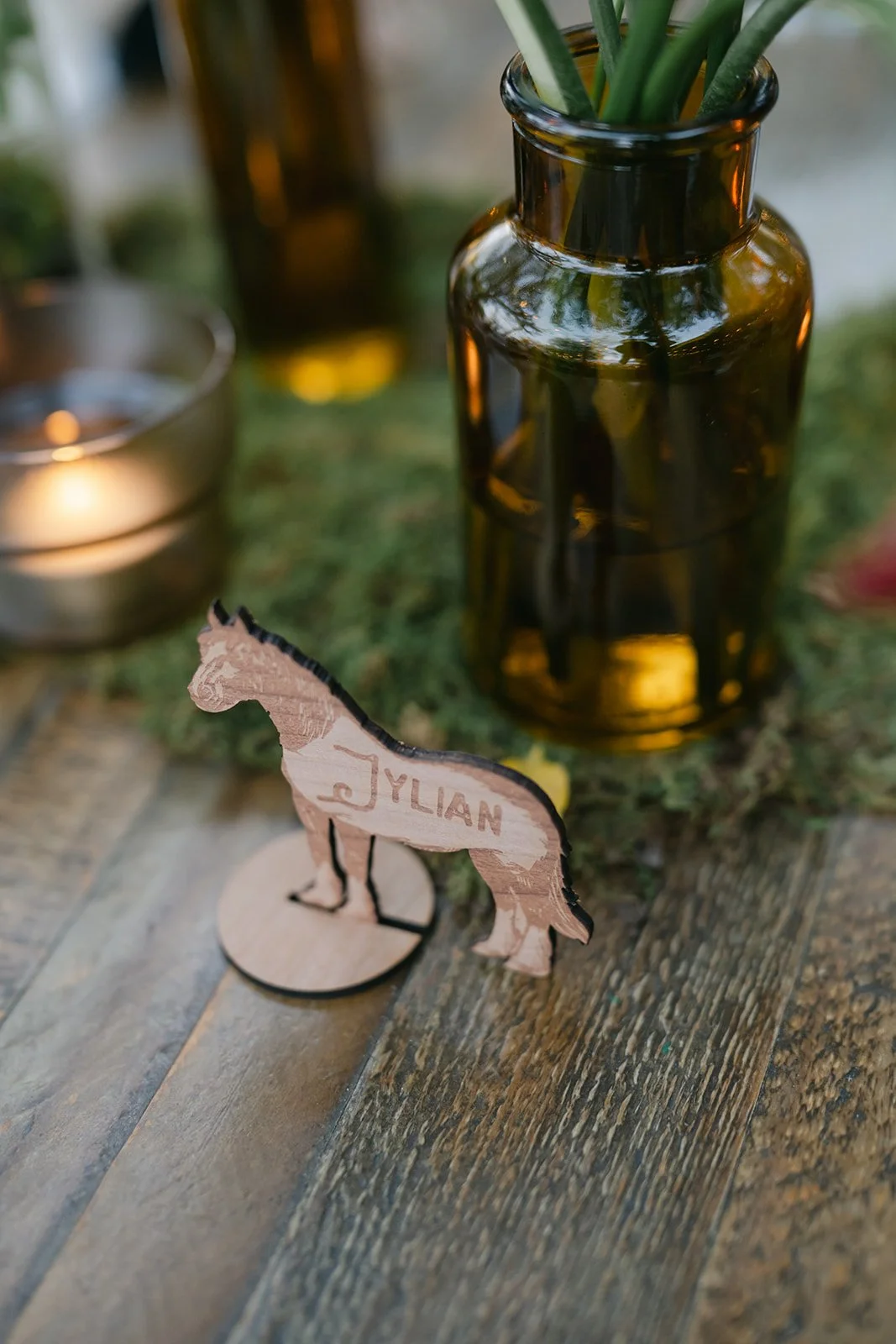A wooden table with a glass jar of green plants, a small metal container, and a wooden cutout of a dog with the name 'JYLIAN' written on it. The photo is taken outdoors with natural lighting.