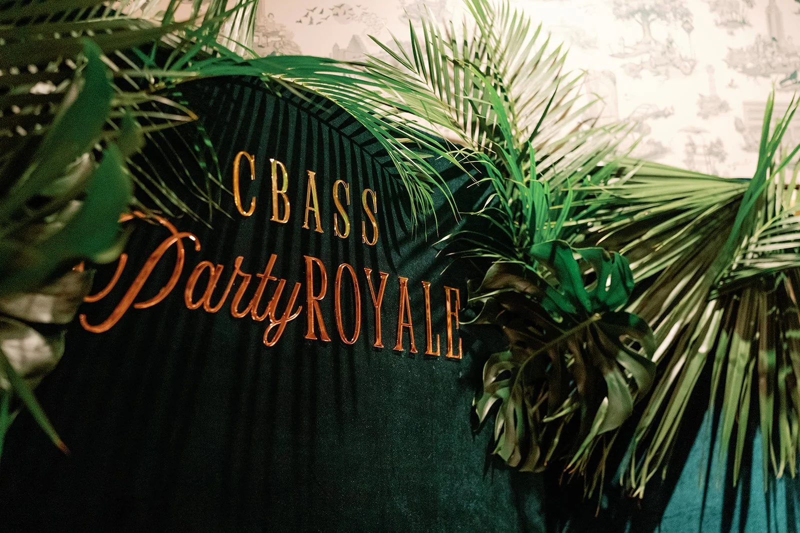 A black sign with gold and copper lettering that reads "C BASS Party ROYALE" is surrounded by various large green tropical leaves, with a light-colored wall in the background.