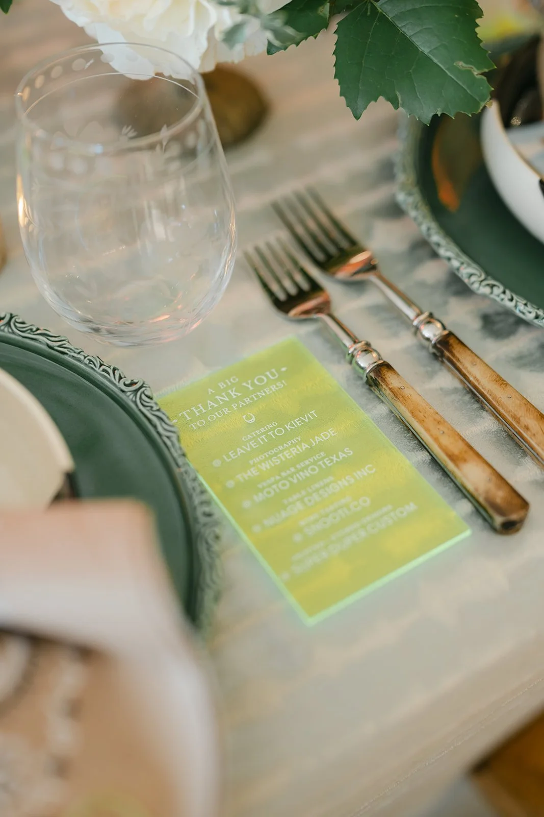 Table setting with a wine glass, two forks with wooden handles, green and white plates, and a yellow thank you card on a marble table.