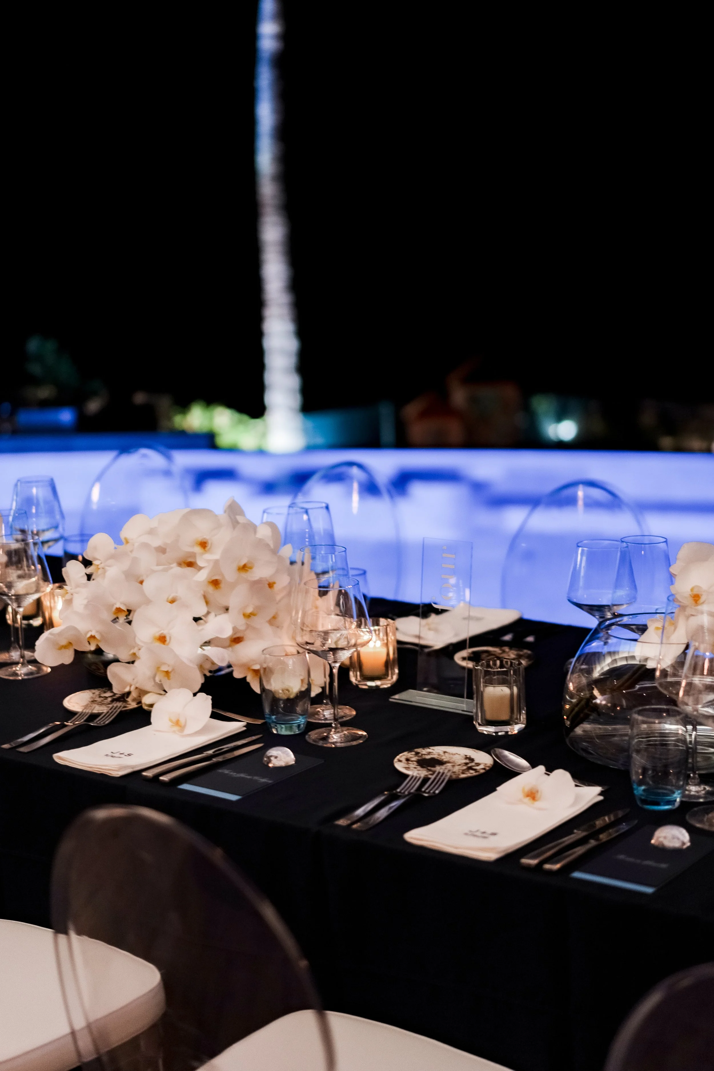 A nighttime outdoor event with a table decorated with white orchids, candles, glasses, silverware, and place settings, with city lights and a tall illuminated building in the background.