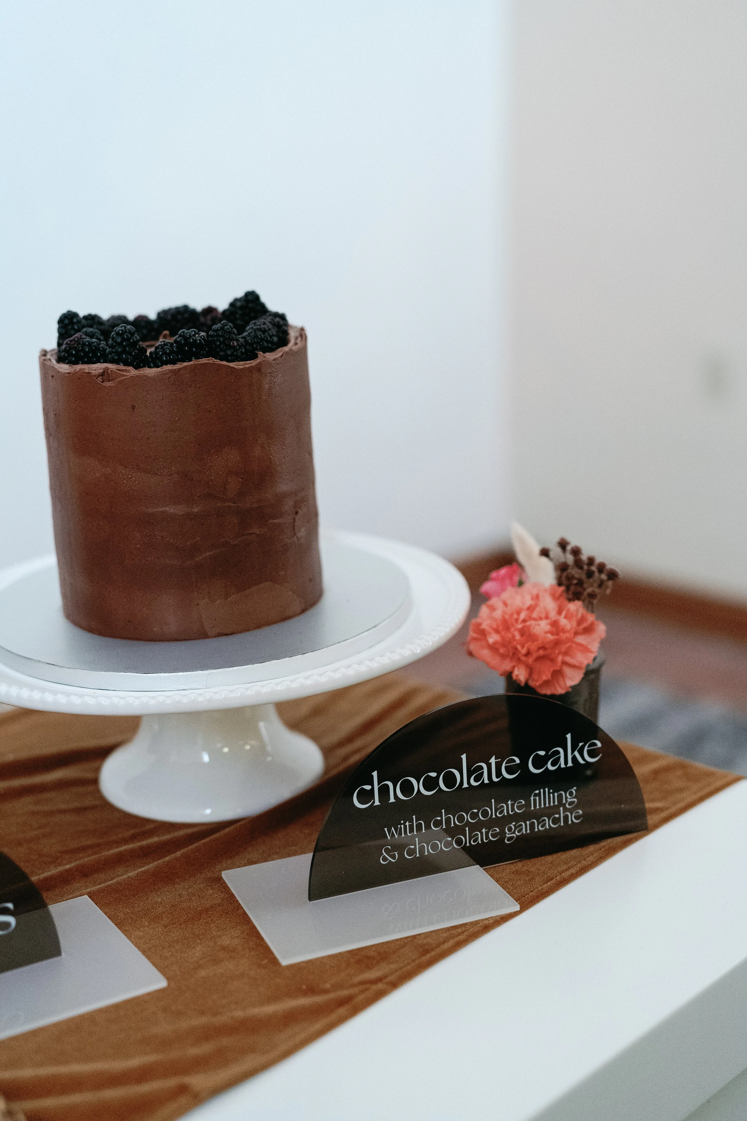 A chocolate cake topped with blackberries on a white cake stand, with a sign reading "Chocolate cake with chocolate filling & chocolate ganache" in front of it, placed on a wooden surface with a pink flower ornament nearby.