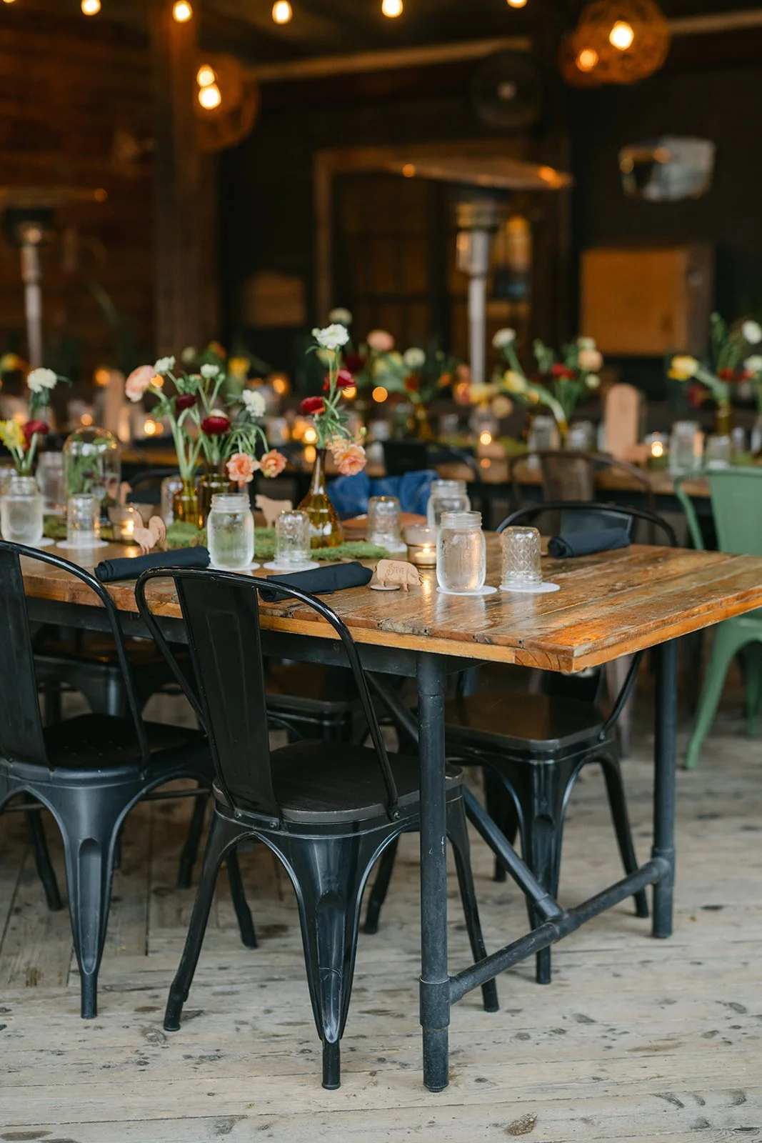 A rustic indoor dining setup with a wooden table decorated with flowers in vases, mason jars filled with water, and candles. The background features dim lighting and additional tables with similar decor.