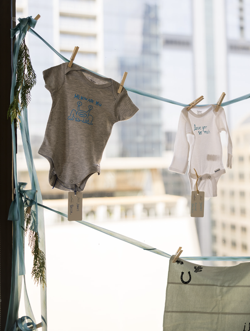 Baby clothes hanging on a clothesline indoors, with tags attached, in front of large windows with city buildings in the background.