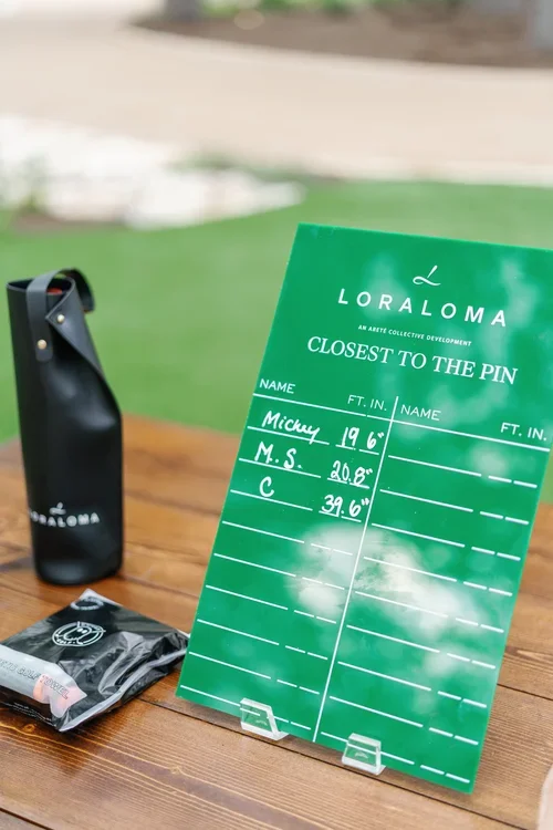 A green outdoor scoreboard from Loraloma with golf distances, placed on a wooden table with a black water bottle and a black packet next to it. Background shows a blurred grassy area.