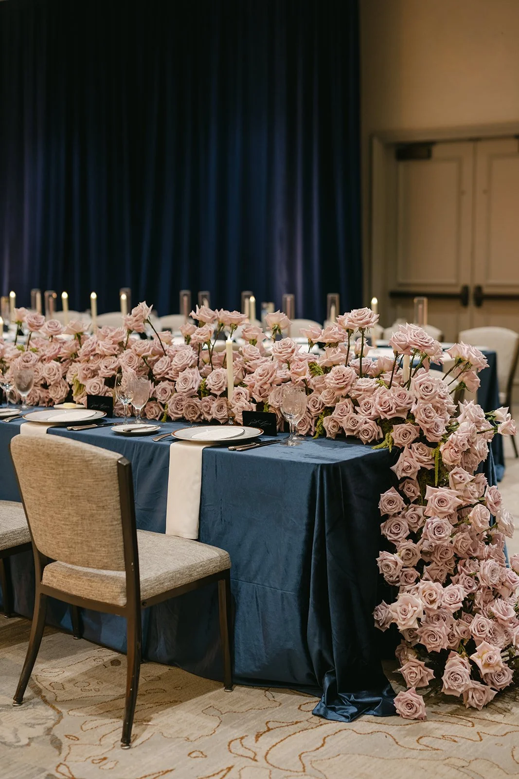 A formal dining table decorated with a large arrangement of pink roses, candles, and elegant tableware, set for a special event or celebration.