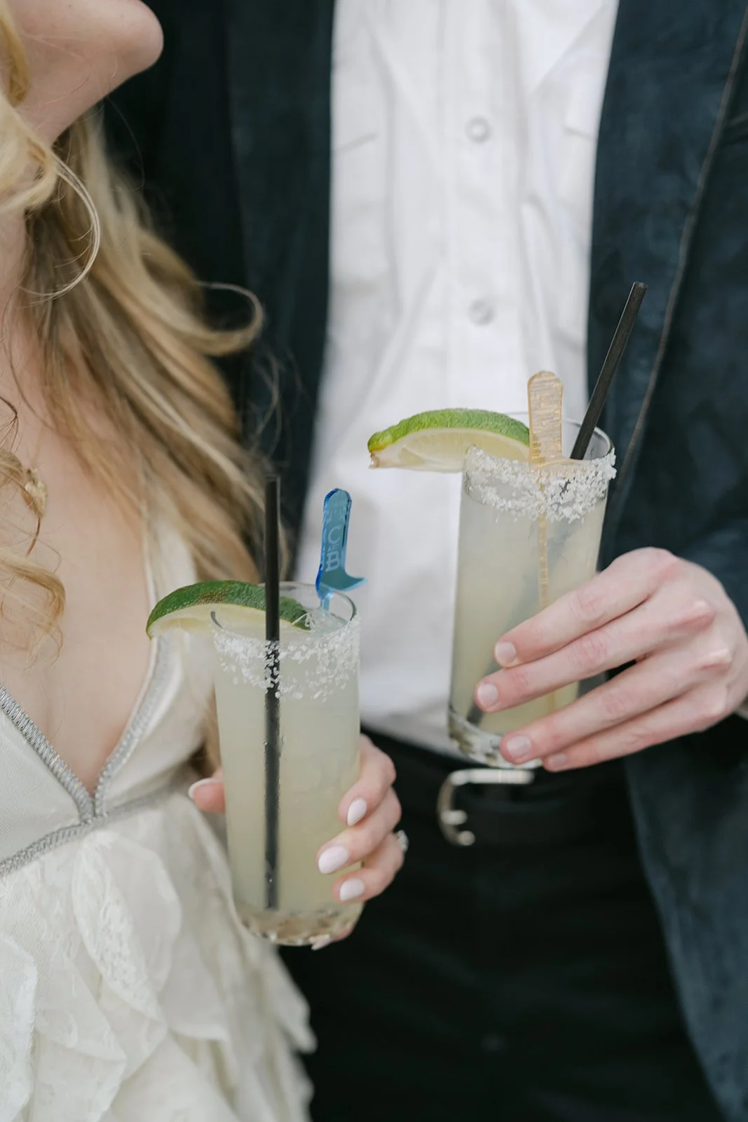 Two people holding mojito cocktails garnished with lime slices, salt rimming the glasses, and plastic stirrers, wearing casual and formal attire.