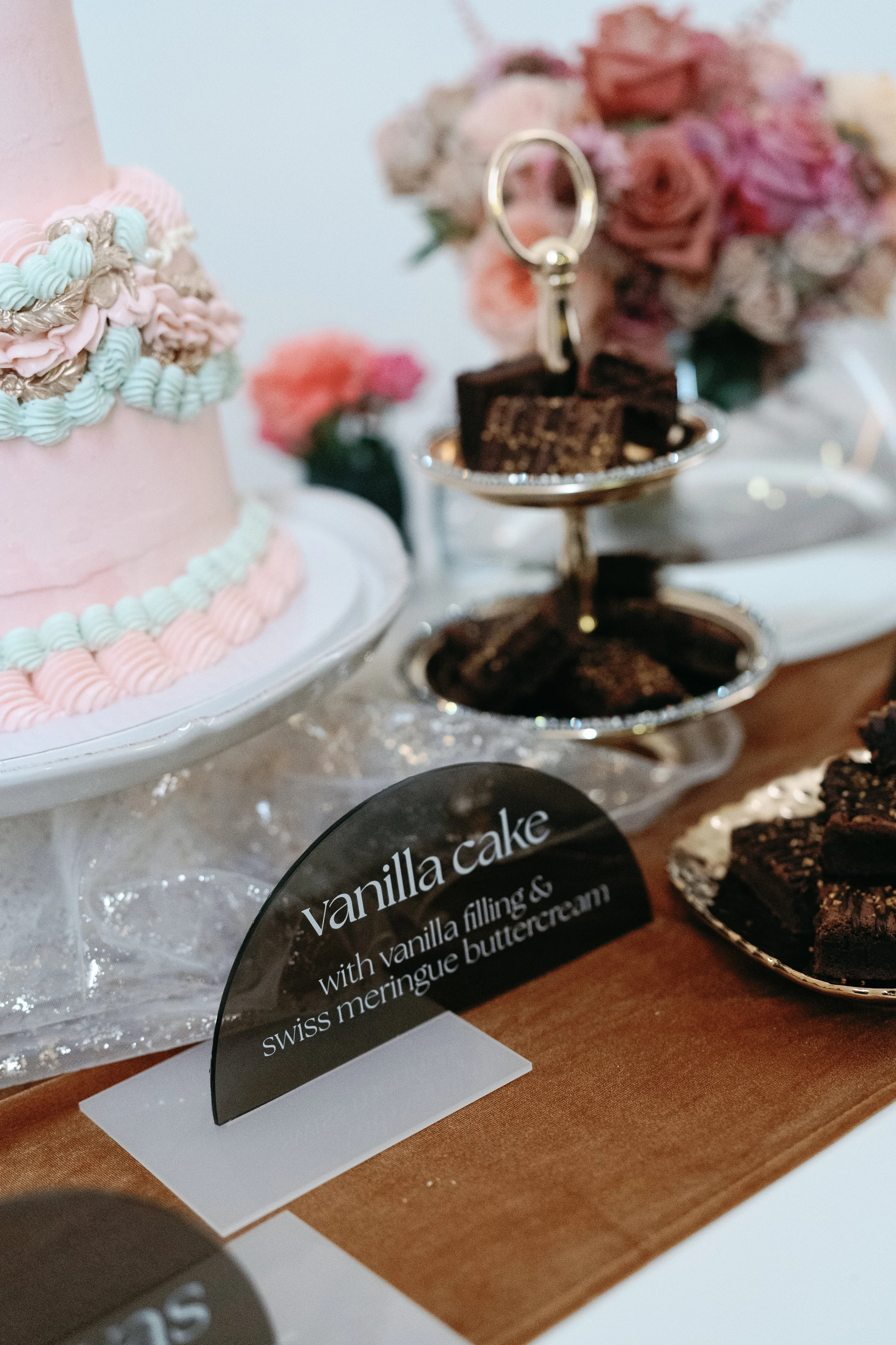 A display of desserts at a party, including a pink cake decorated with pastel-colored icing and a bouquet of pink roses. There is a small dish of chocolate brownies and a sign that reads 'Vanilla cake with vanilla filling & swiss meringue buttercream