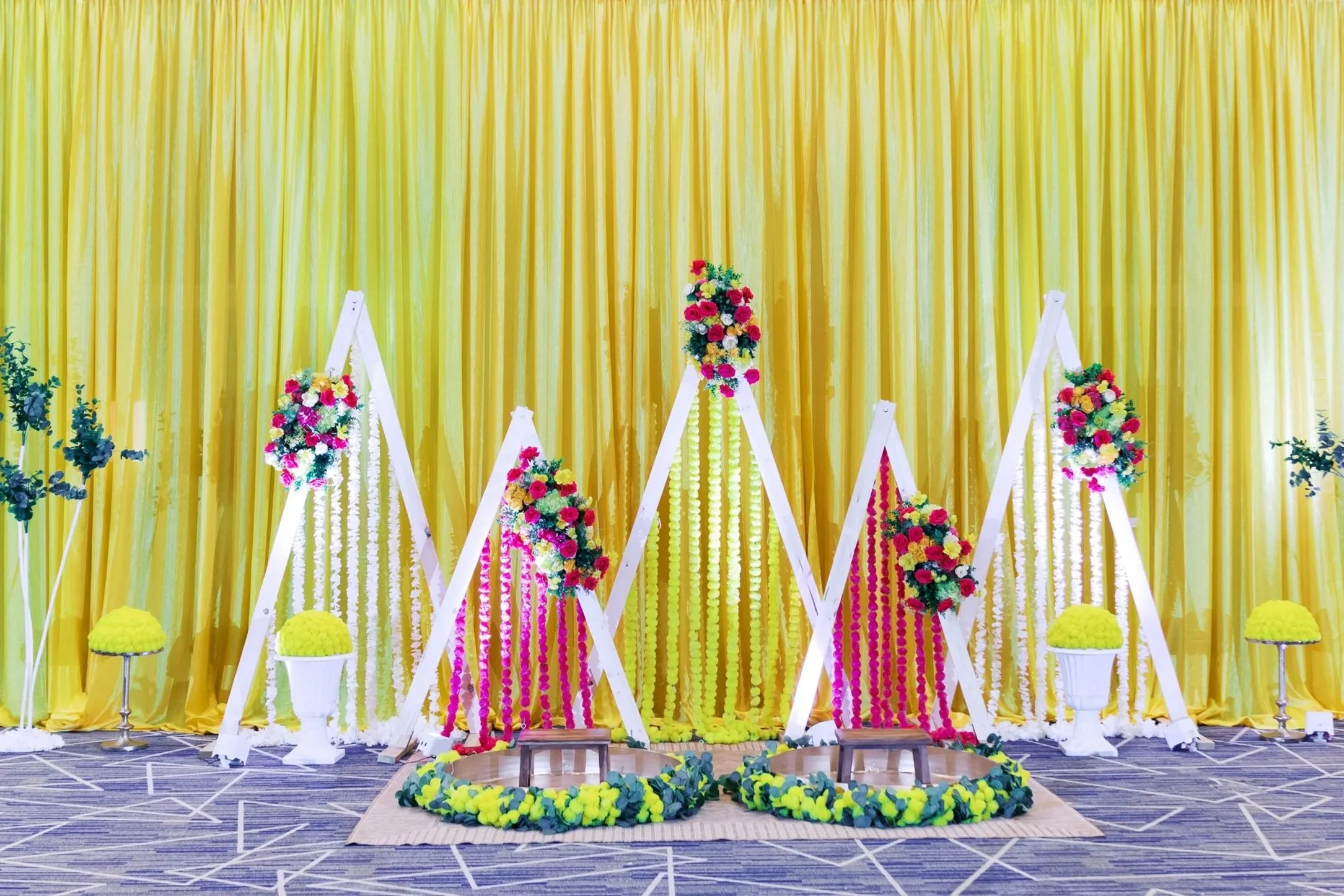 Vibrant Indian wedding stage decoration with multi-colored drapes, embroidered umbrellas, a gold ornate sofa, decorative pillows, potted plants, and illuminated trees.