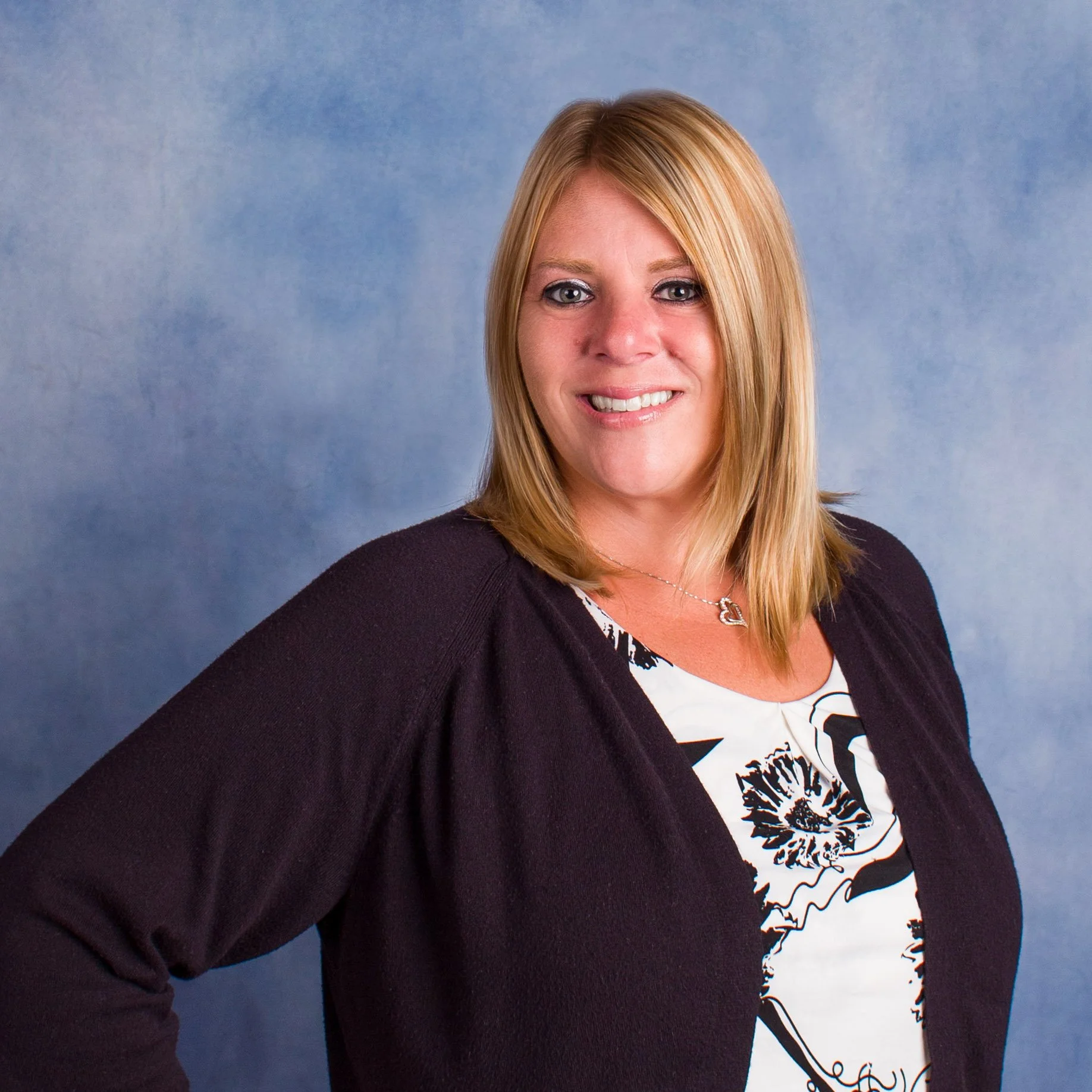 Portrait of a woman with short, blonde hair wearing a black top and a necklace, smiling against a green background.