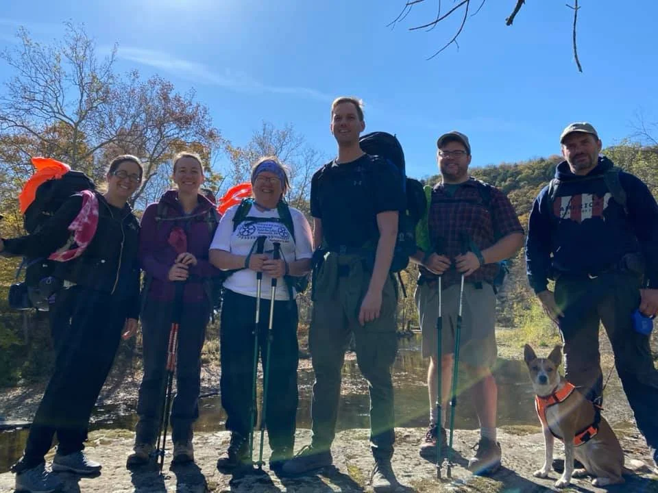 Group of six people and one dog hiking outdoors near a river, with trees and a blue sky in the background.