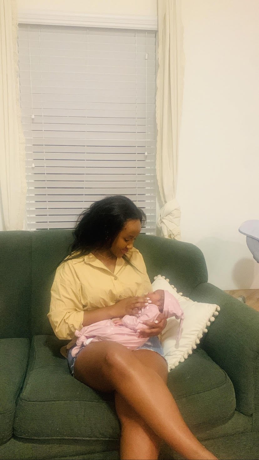 A woman sitting on a green couch holding a small baby wrapped in pink clothing, in a living room with cream curtains and white blinds.
